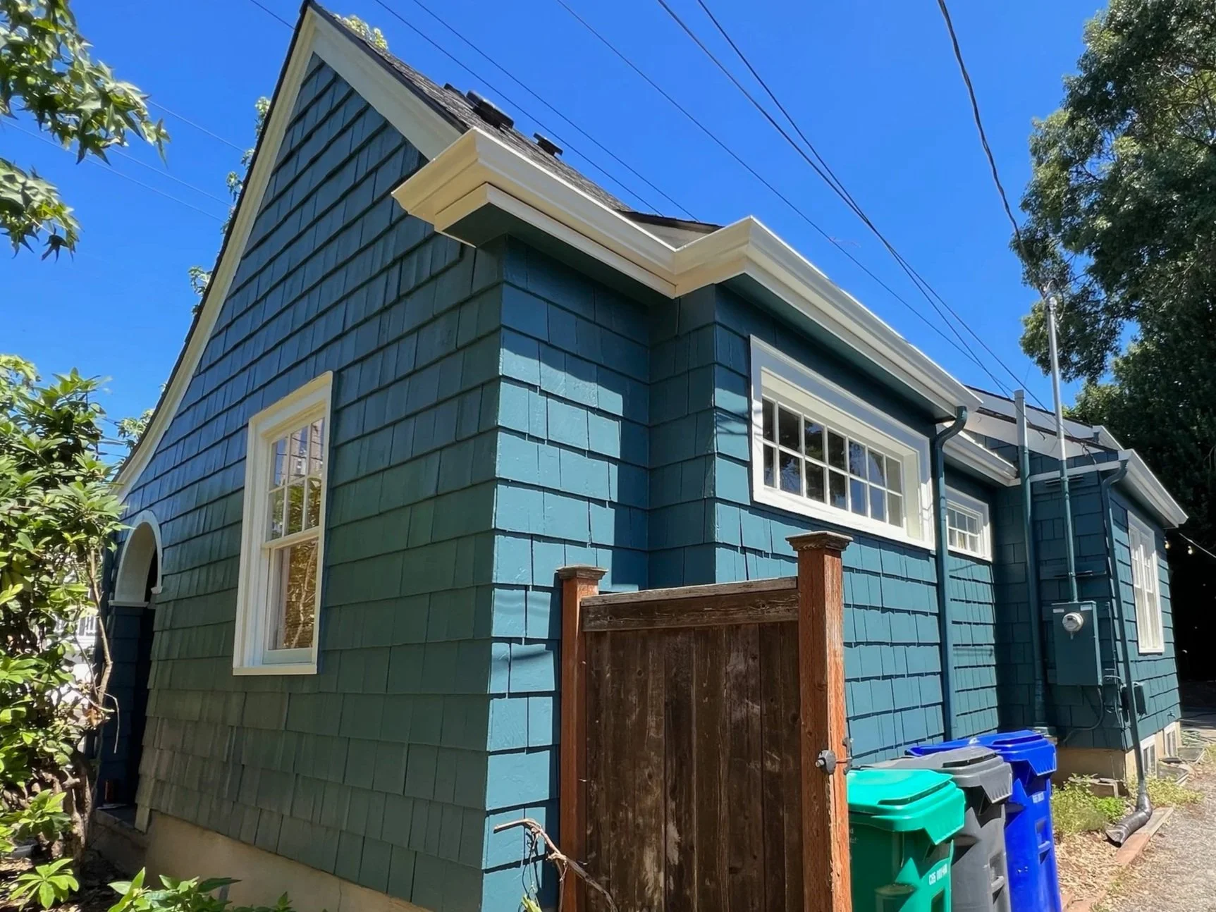 Backyard view of a blue house with white trim, a wooden fence, and four garbage cans on a small cement platform. There are tall trees and a clear blue sky in the background.