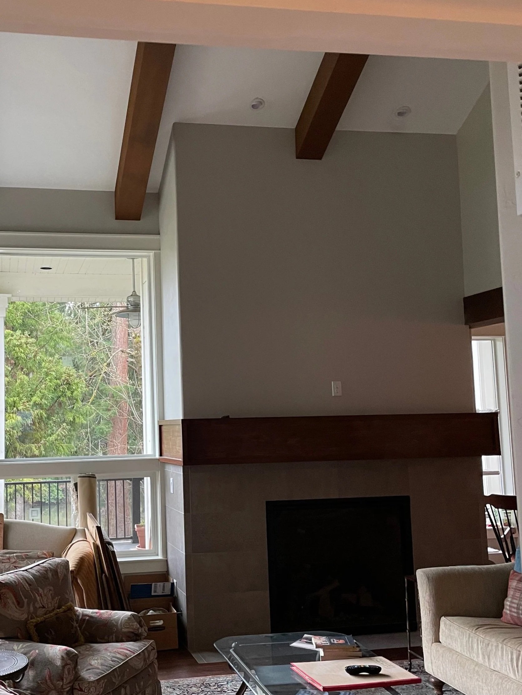 Living room with large windows, beige and floral upholstered furniture, a glass coffee table, a fireplace, and various items like books and boxes scattered around.