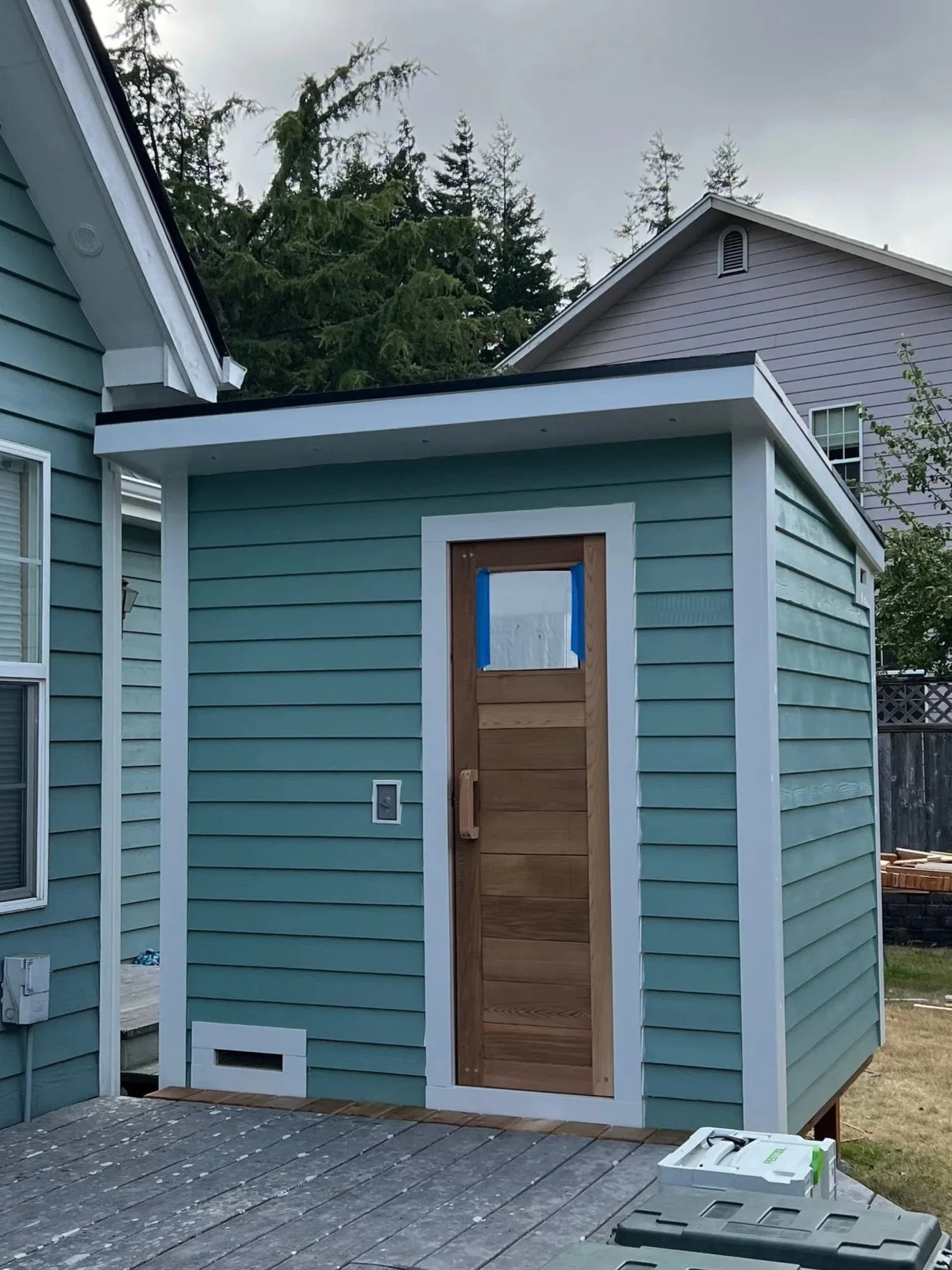 Backyard with a wooden deck and a blue shed under cloudy sky, new wooden door, and construction materials.