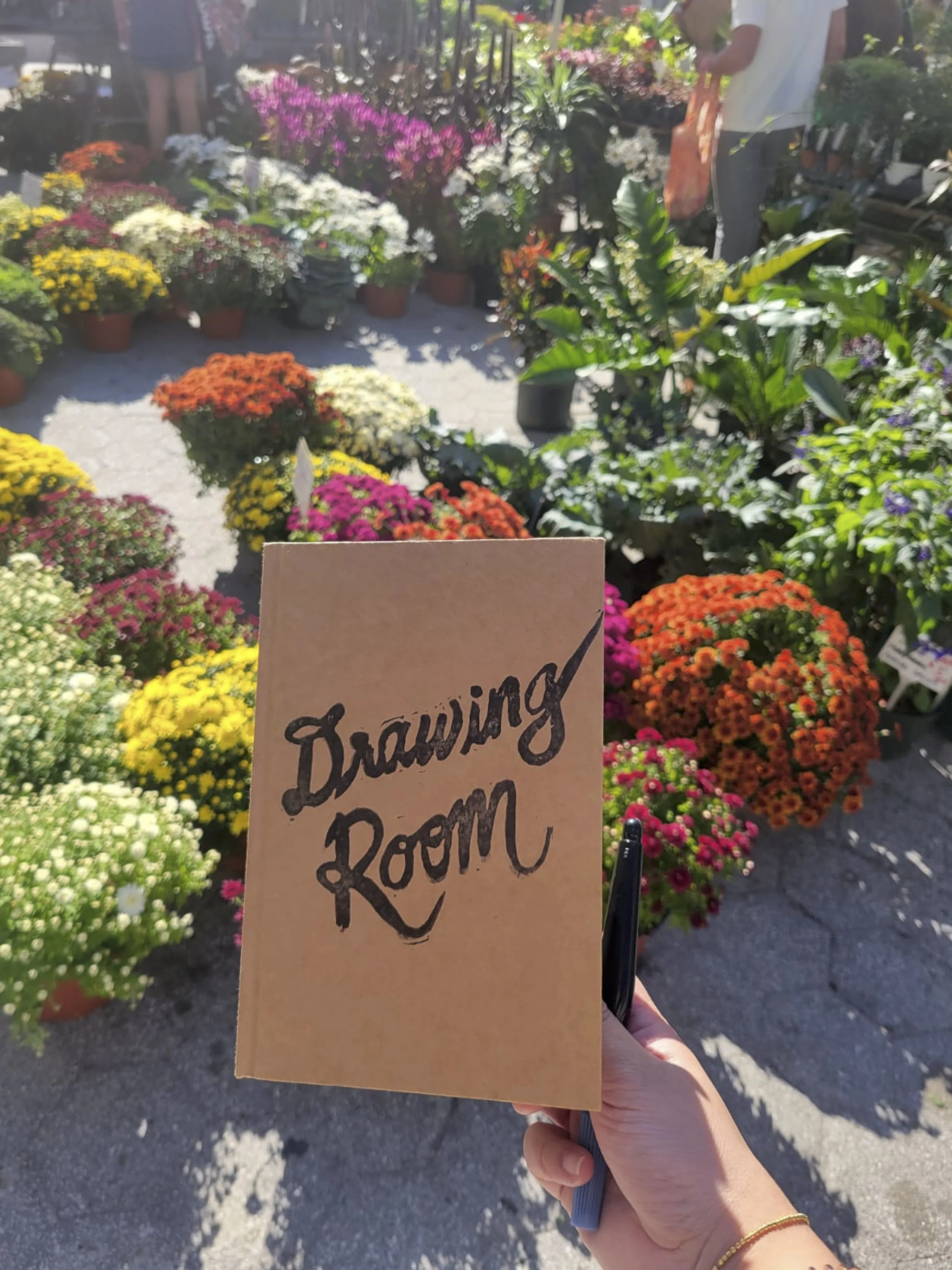 A person holding a paper that says 'Drawing Room' in front of a colorful flower nursery with potted plants and flowers in full bloom.
