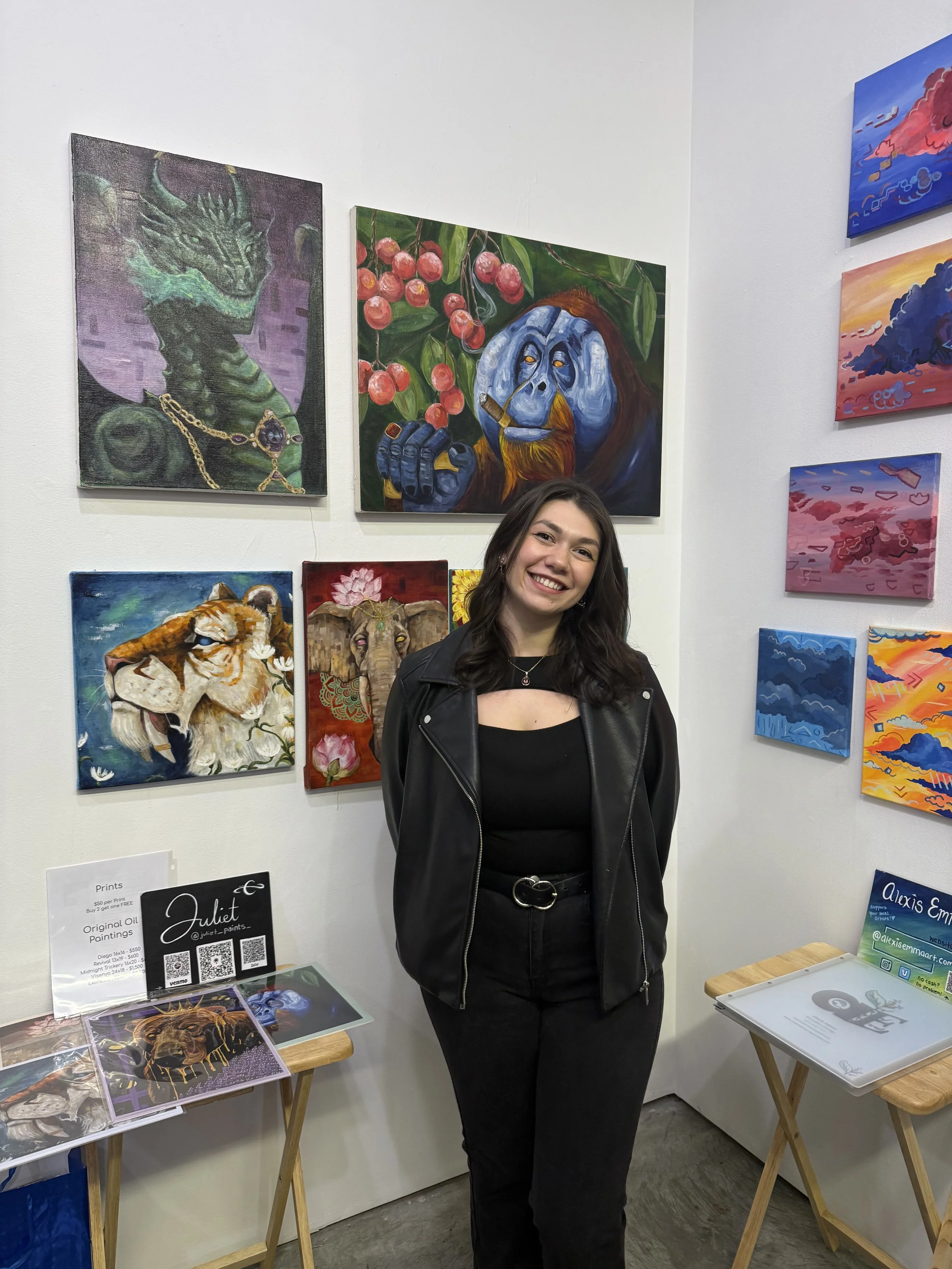 A woman with shoulder-length dark hair, wearing a black outfit and a black leather jacket, smiling and standing in front of a display of colorful paintings of animals, including a dragon, gorilla, tiger, and elephant, at an art gallery.