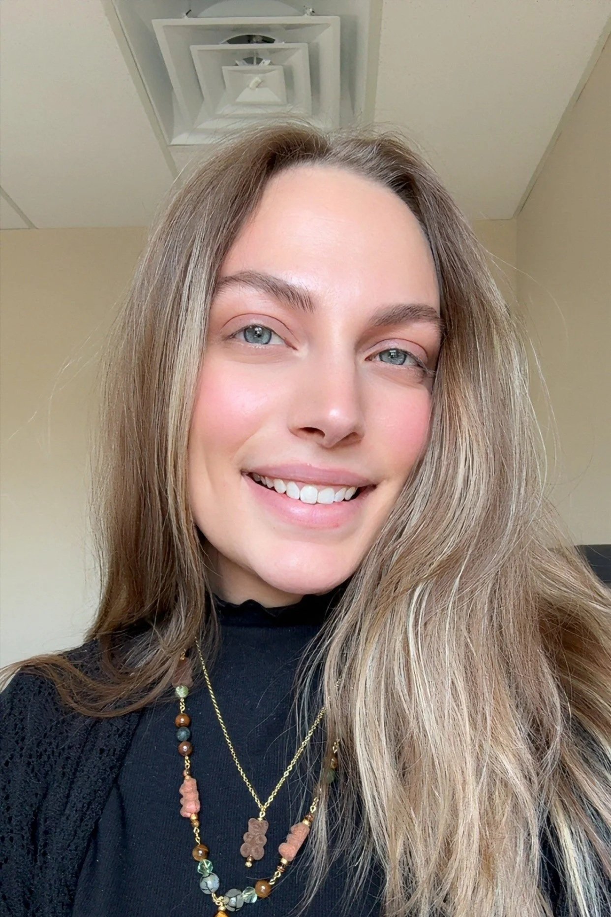 Close-up photo of a young woman with long, wavy blonde hair, blue eyes, smiling, wearing black top and layered beaded necklaces.