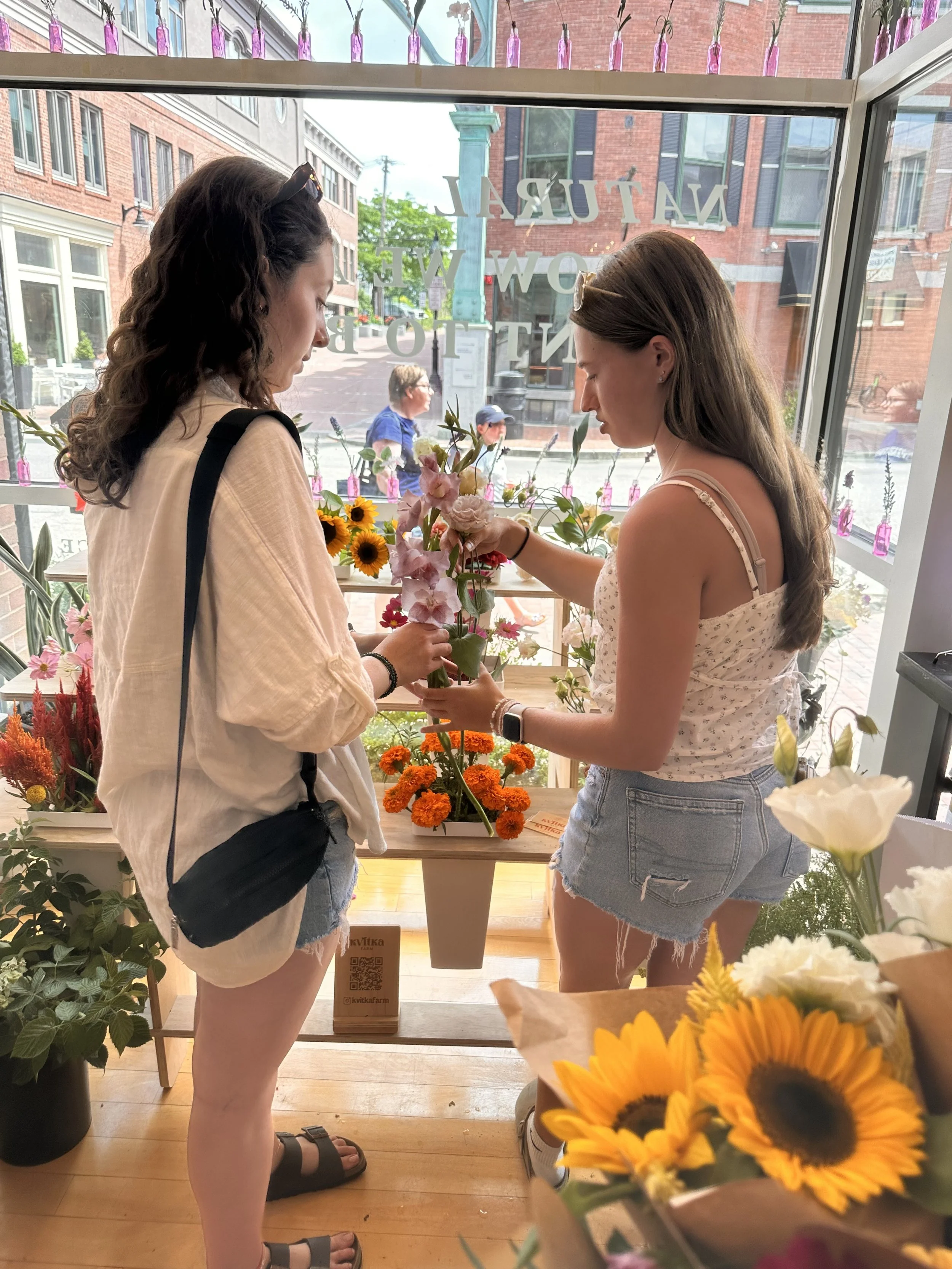 Two young women shopping for flowers inside a flower shop. They are standing near a wooden table with various colorful flowers, including sunflowers and pink blooms. The shop has large windows with city sidewalk view outside. One woman is wearing a white blouse, denim shorts, and sandals, while the other is in a light-colored tank top and shorts, with a black bag over her shoulder.