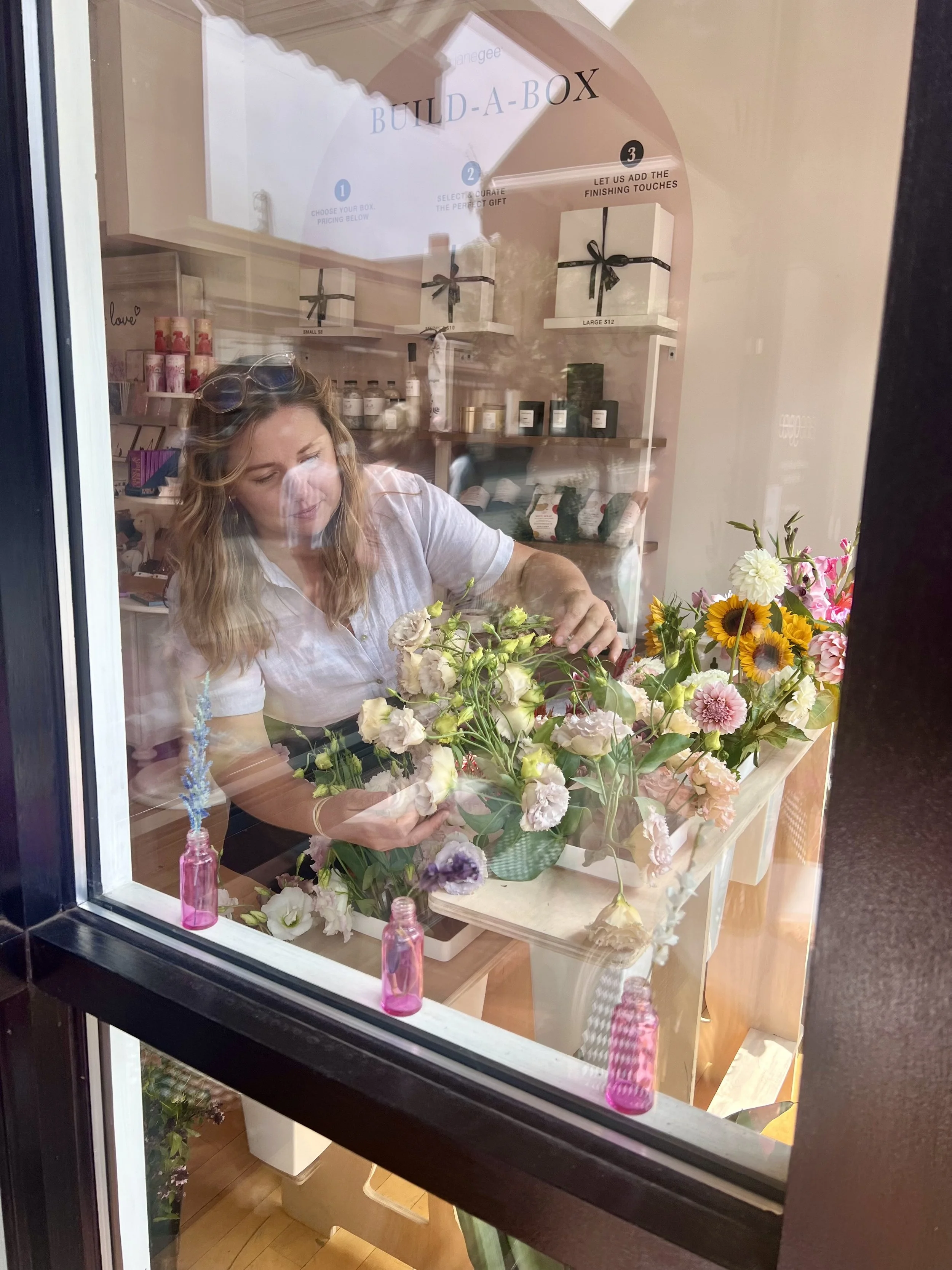 A woman arranging flowers inside a flower shop, seen through a glass window with reflection of the street outside.