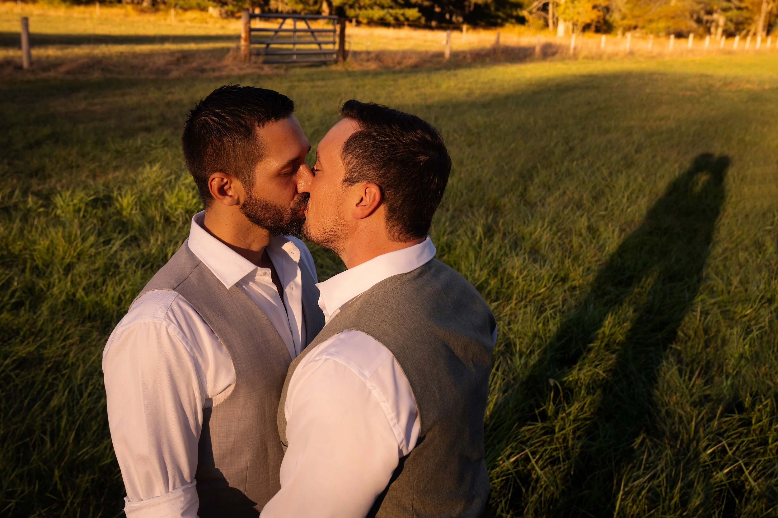 Captured from above during golden hour, two grooms share a kiss surrounded by glowing green grass lit by the setting sun. Their silhouettes stretch behind them, forming two tender shadows meeting in the background. The warm light and elevated view cr