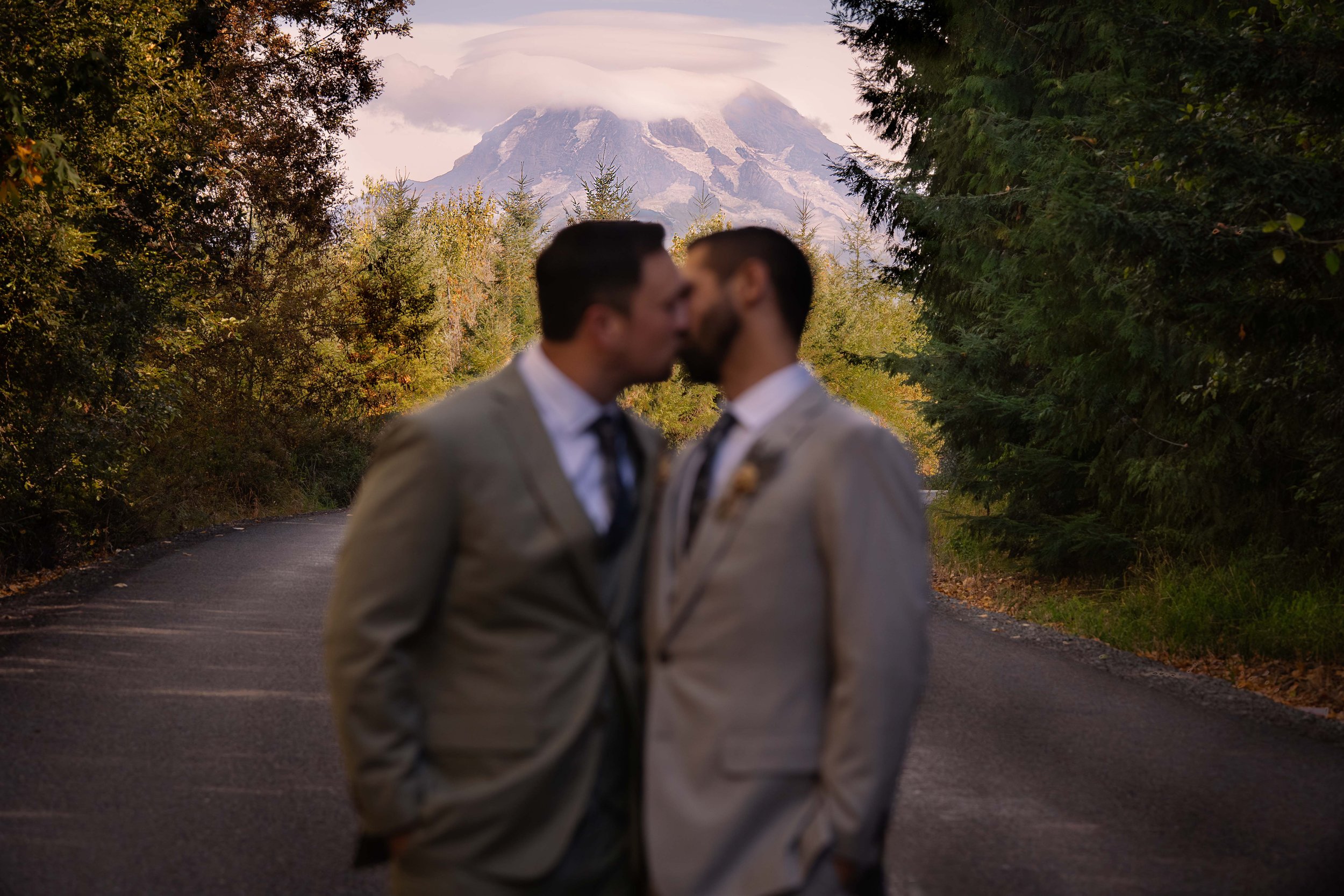 A quiet, emotional moment between two grooms on their wedding day - sharing a kiss as Mount Rainer stands tall and clear behind them. The photo has dark, moody, and romantic feel, with the couple softly blurred in the foreground and the mountain in s