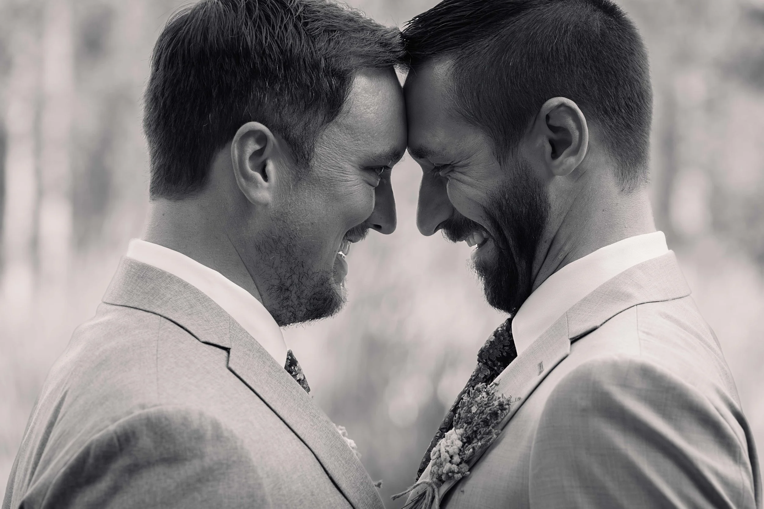 Two grooms stand close, foreheads gently touching, sharing a soft laugh just after their wedding ceremony. Captured in timeless black and white, their joy and connection shine through as the background fades into a romantic blur. A candid, heartfelt 