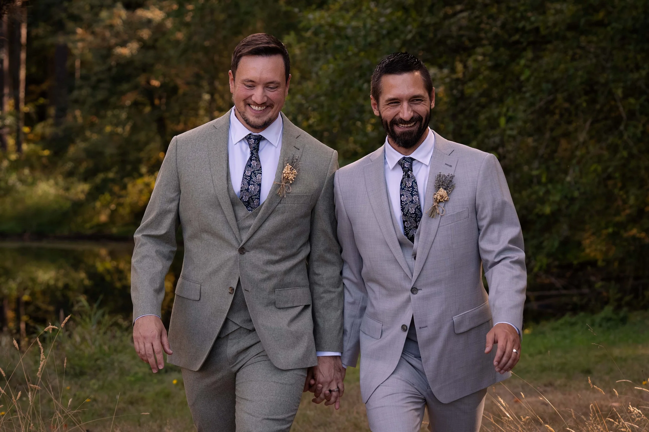 Two grooms walk hand in hand towards the camera, laughing together in a moment of pure happiness. Behind them, sunlight filters through the trees and leaves, casting a warm, golden glow over the scene. The sparkle of water shimmers softly in the back