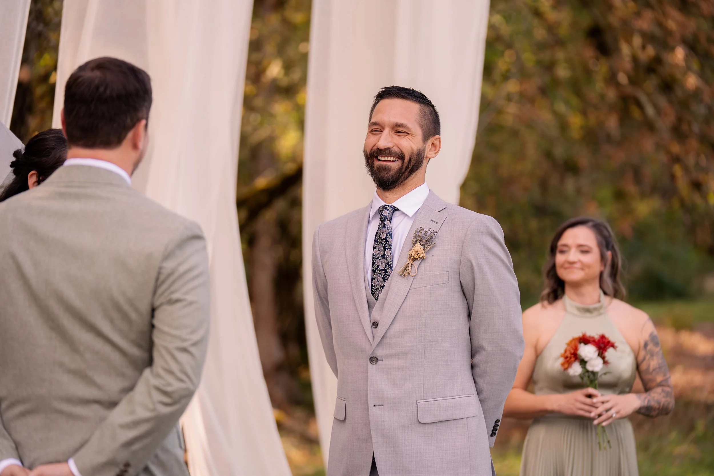 During their fall wedding ceremony, one groom is caught mid-laughter - giggling at something sweet that was said - his face glowing with love and joy. Behind him, soft white linens sway gentle in the autumn breeze, and the background fades into a dre