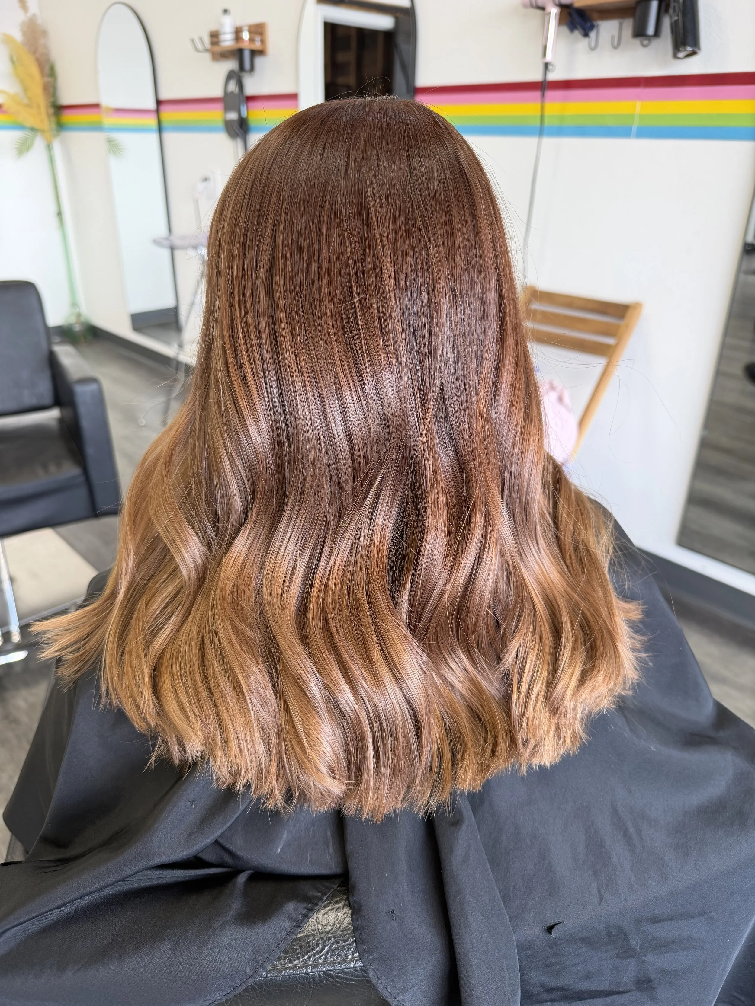 A person with long, wavy, reddish-brown hair sitting in a salon chair, with salon equipment visible in the background.