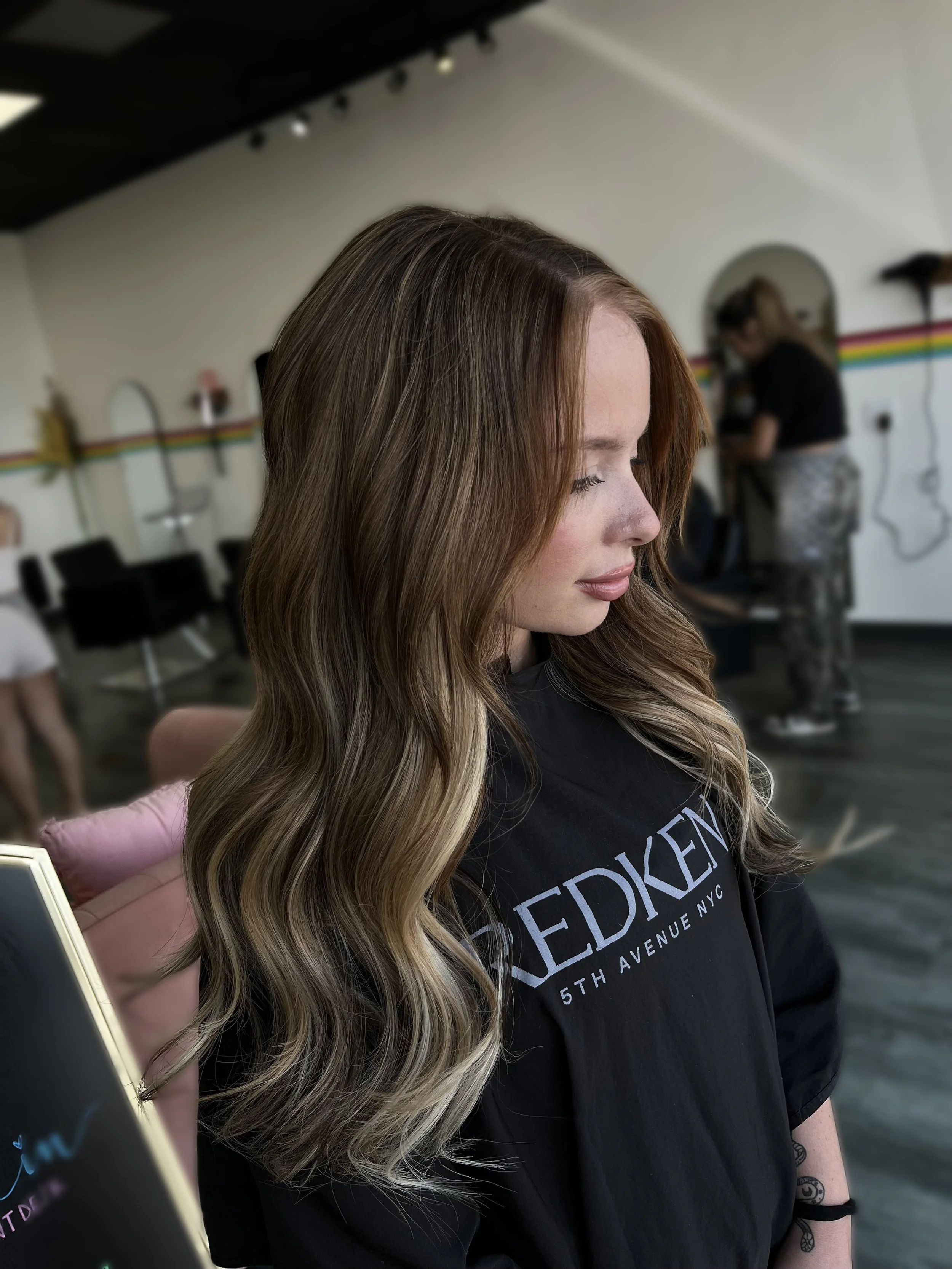 A woman with long wavy hair looking down, wearing a black T-shirt with white text, in a salon with chairs and another person in the background.