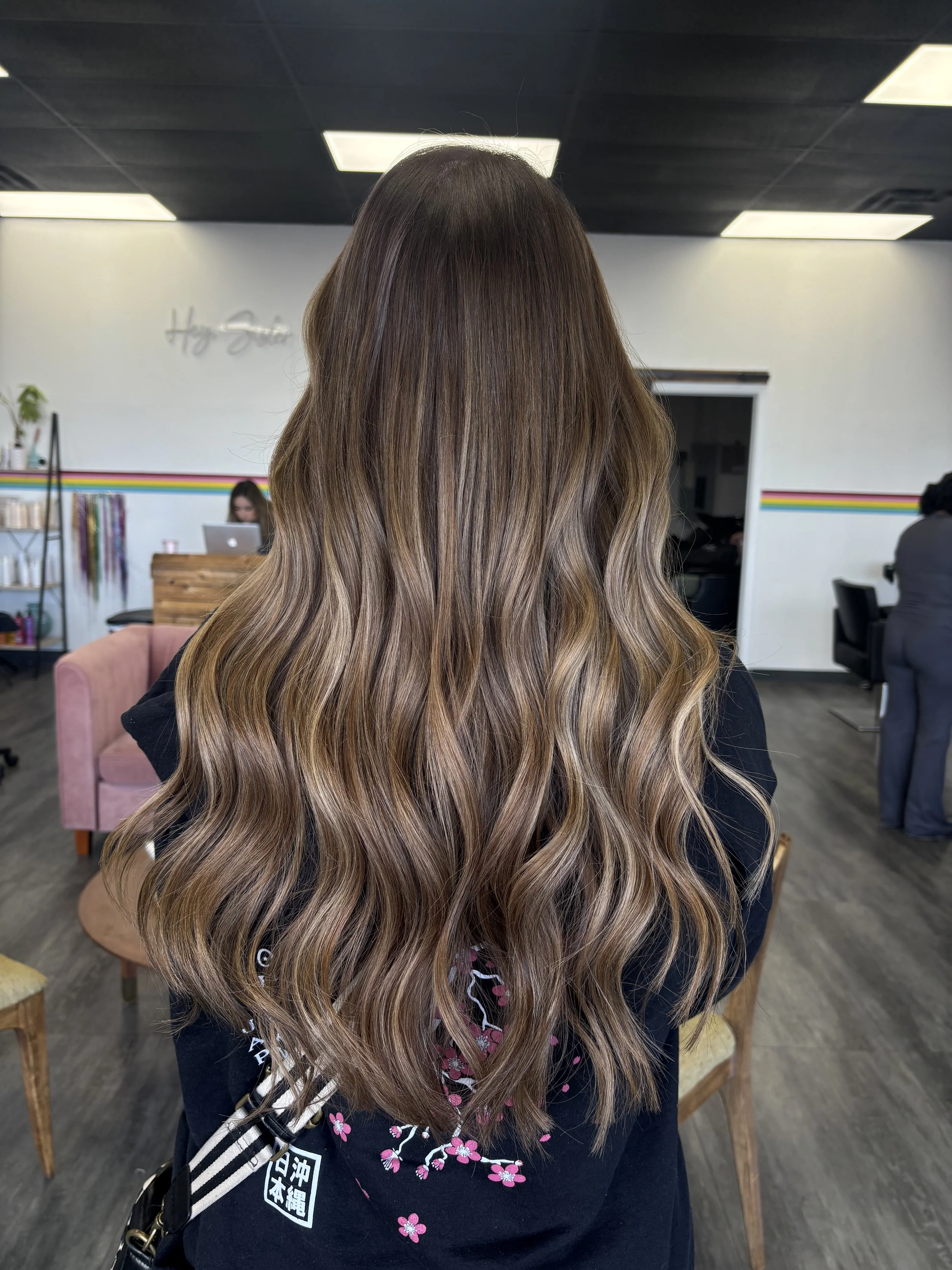 Back view of a woman with long, wavy, ombré hair in shades of brown and blonde, sitting in a salon with white walls, black ceiling tiles, bright rectangular ceiling lights, and salon furniture.