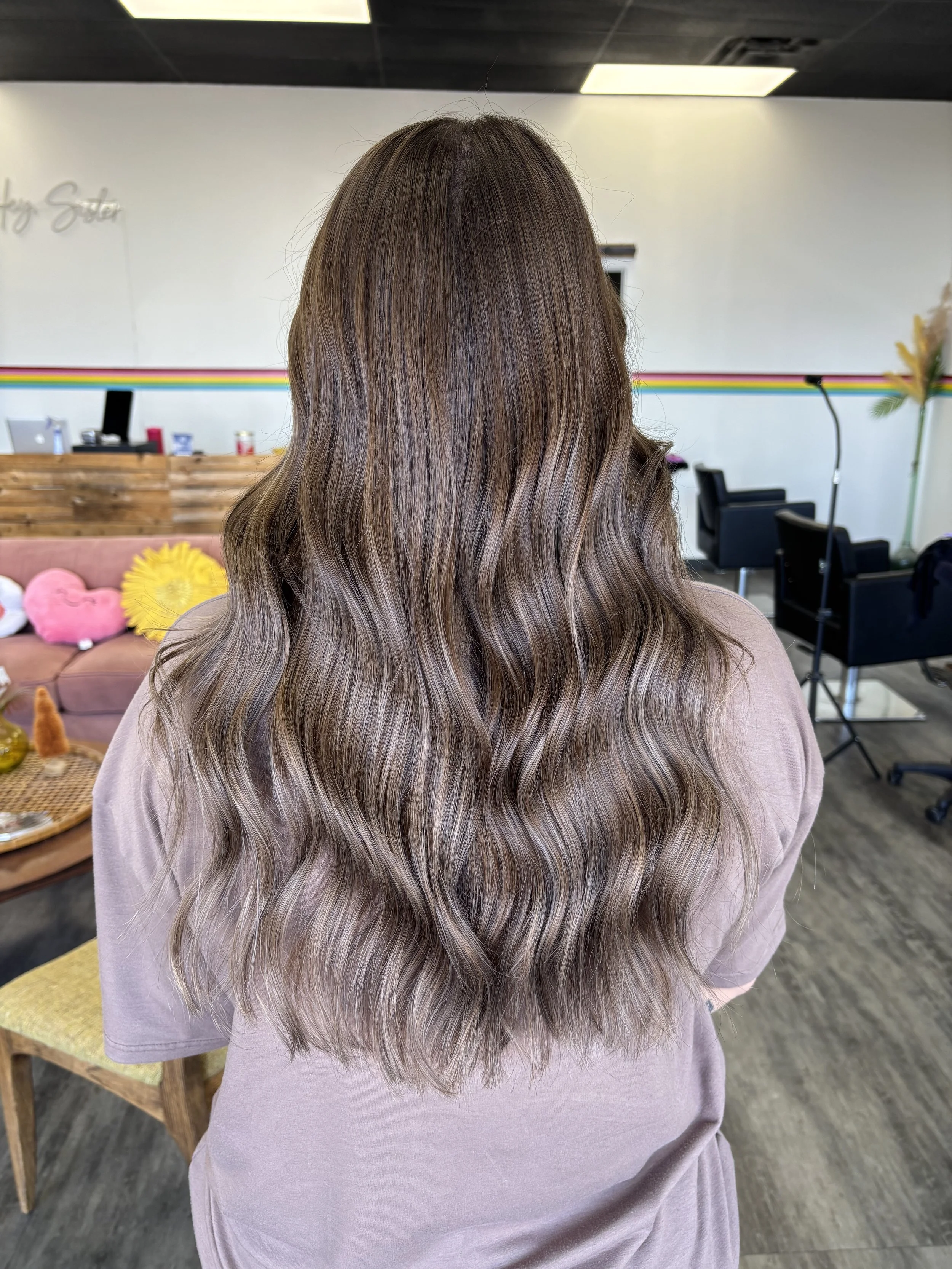 Back view of a woman with long, wavy, brown hair with subtle highlights, wearing a mauve top, in a hair salon with chairs, a reception desk, and colorful decor.