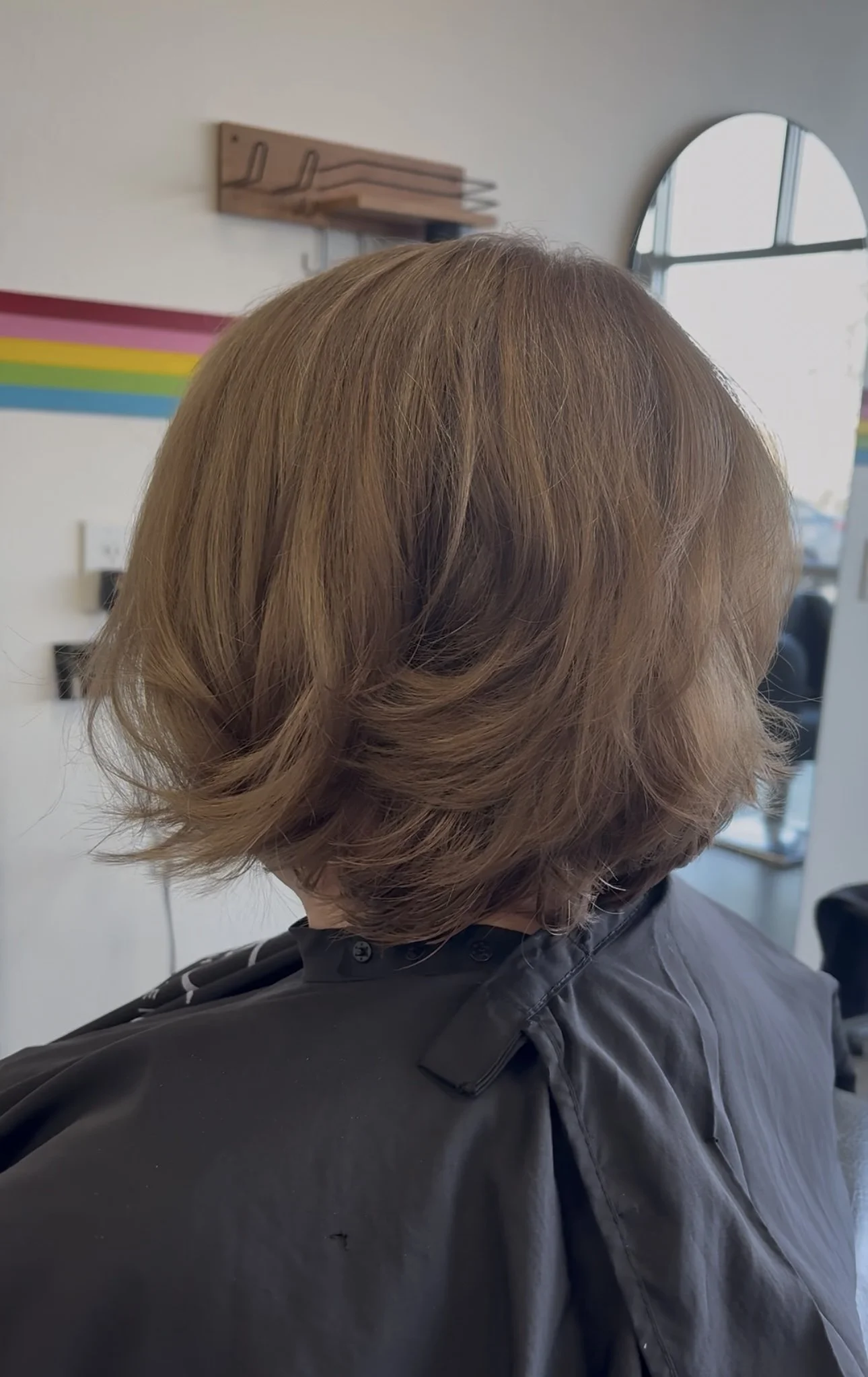 The image shows a woman with shoulder-length, wavy, light brown hair sitting in a salon chair, wearing a black salon cape. The background includes a mirror and a rainbow-colored stripe on the wall.
