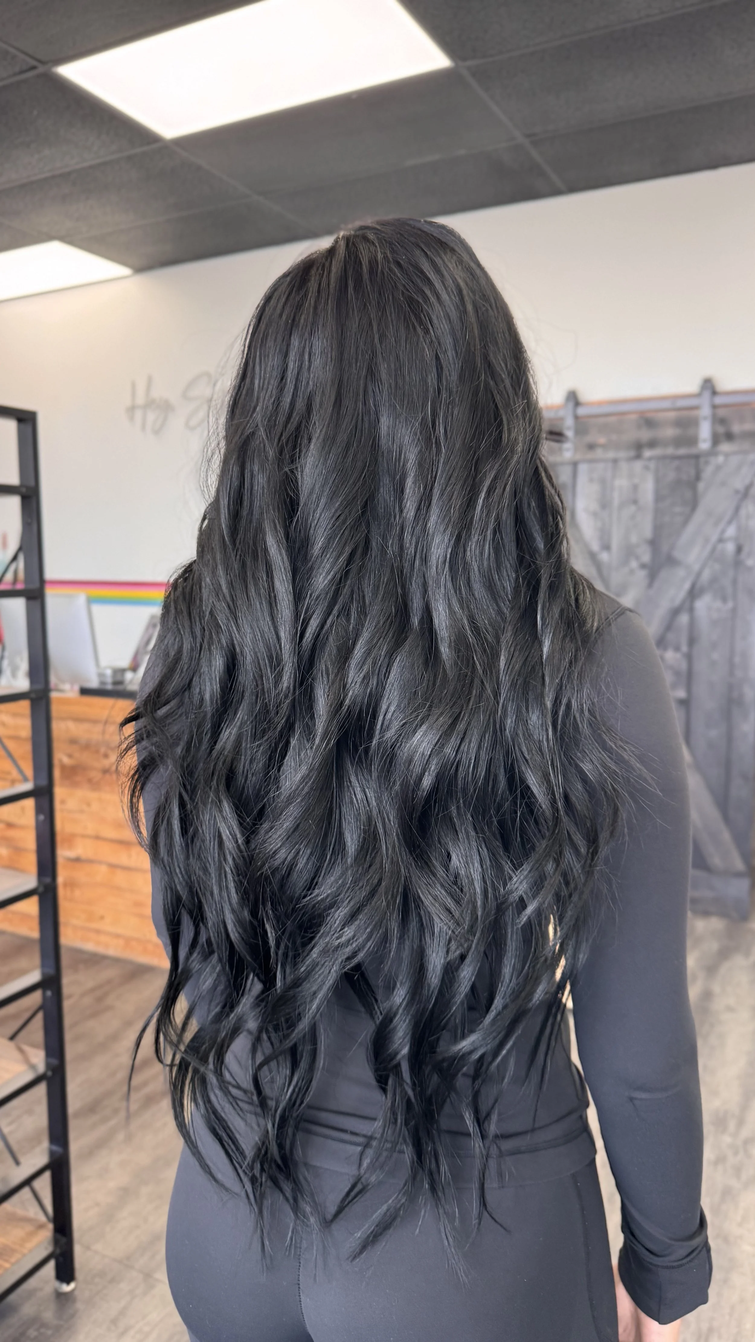 Woman with long, wavy black hair, wearing a black top, standing indoors with a rustic wooden door and modern ceiling lights in the background.