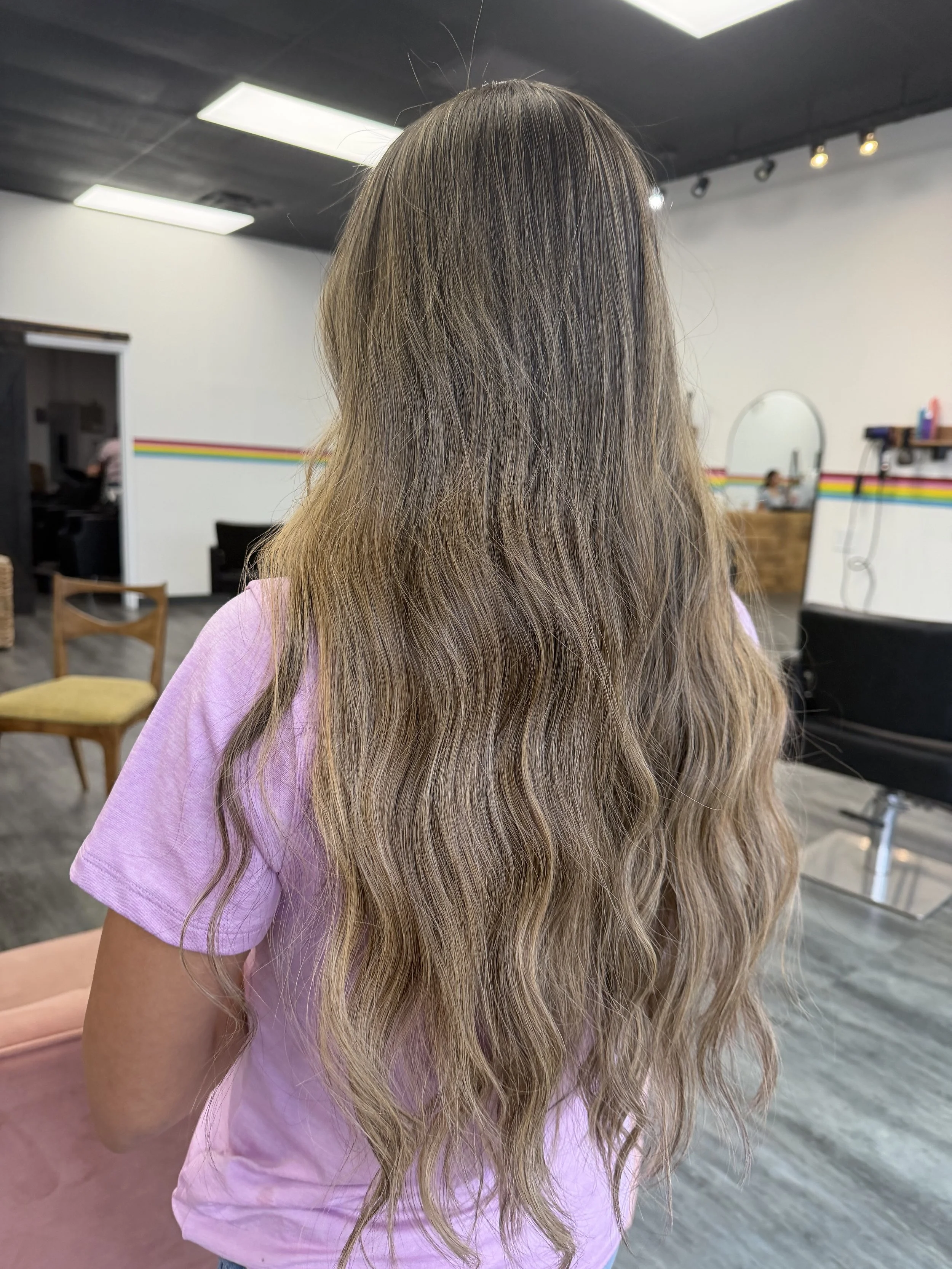 Back view of a woman with long, wavy, blonde hair wearing a pink shirt inside a hair salon.