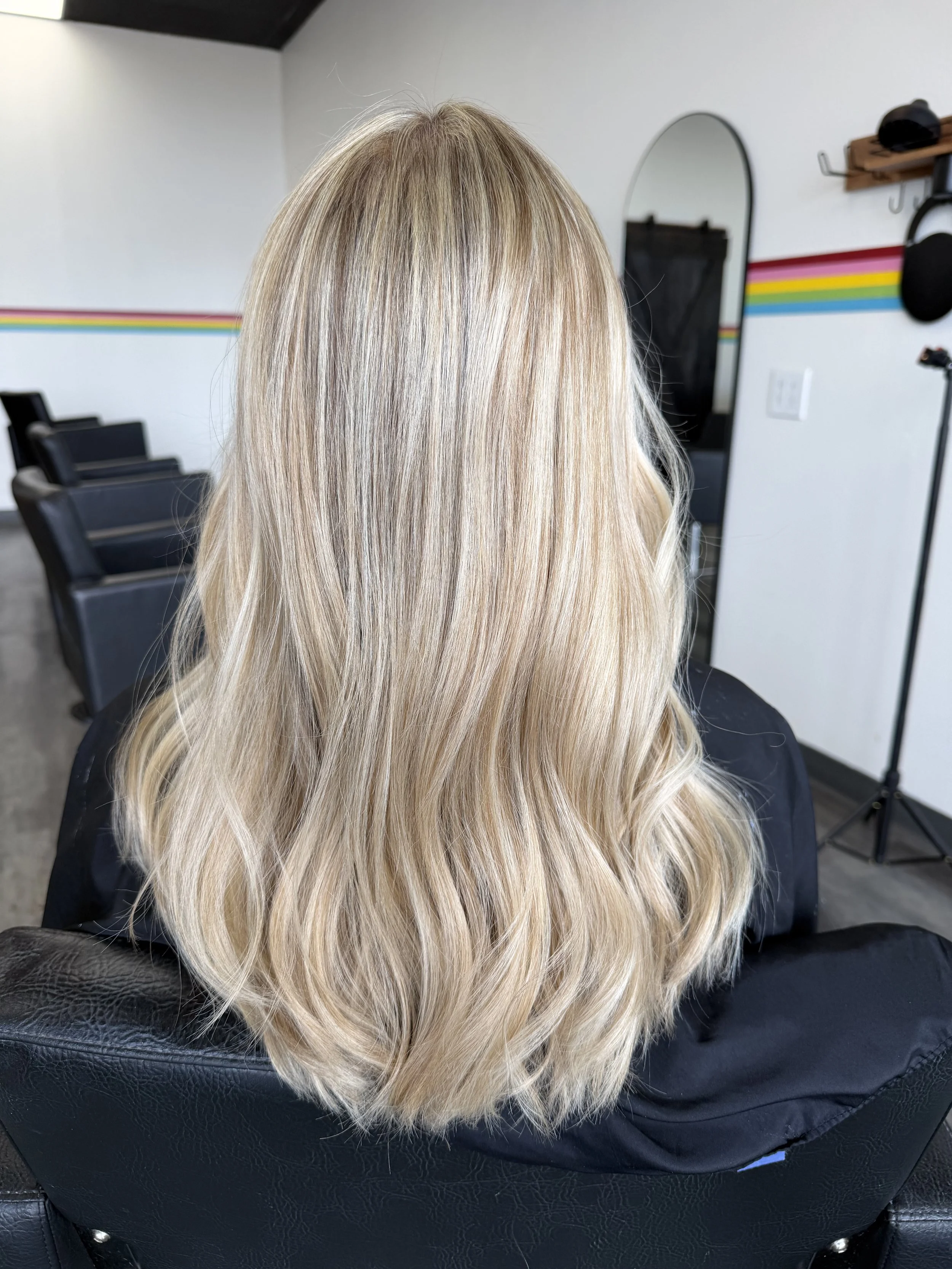 A woman with long, straight, blonde hair sitting in a salon chair, seen from the back, with a mirror and salon tools in the background.