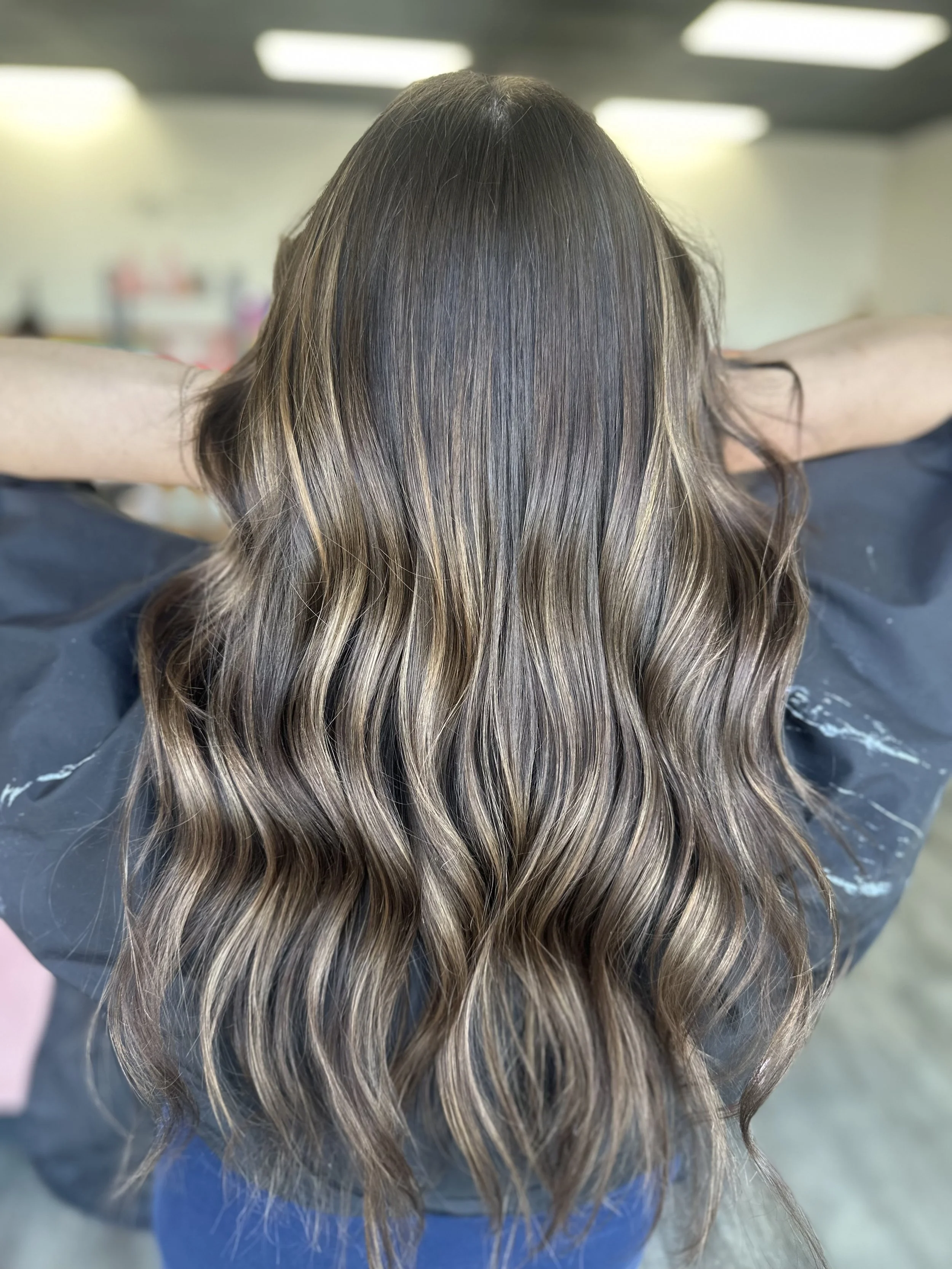 Back view of a woman with long, wavy hair that has blonde highlights and darker roots, sitting with her arms behind her head in a salon or similar setting.