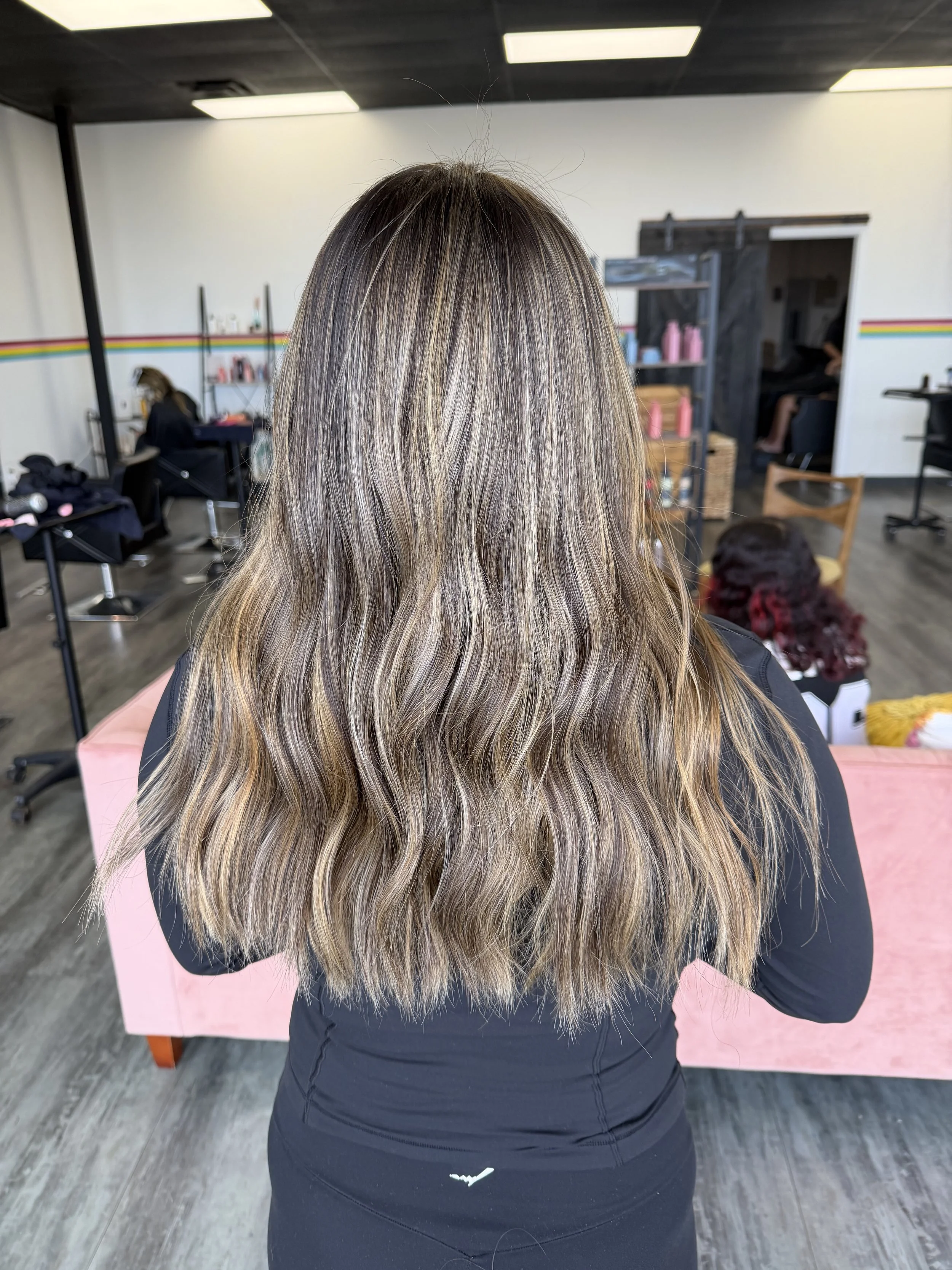 Back view of a woman with long, wavy hair with blonde highlights sitting in a salon, facing away from the camera, with hair styled and colored.