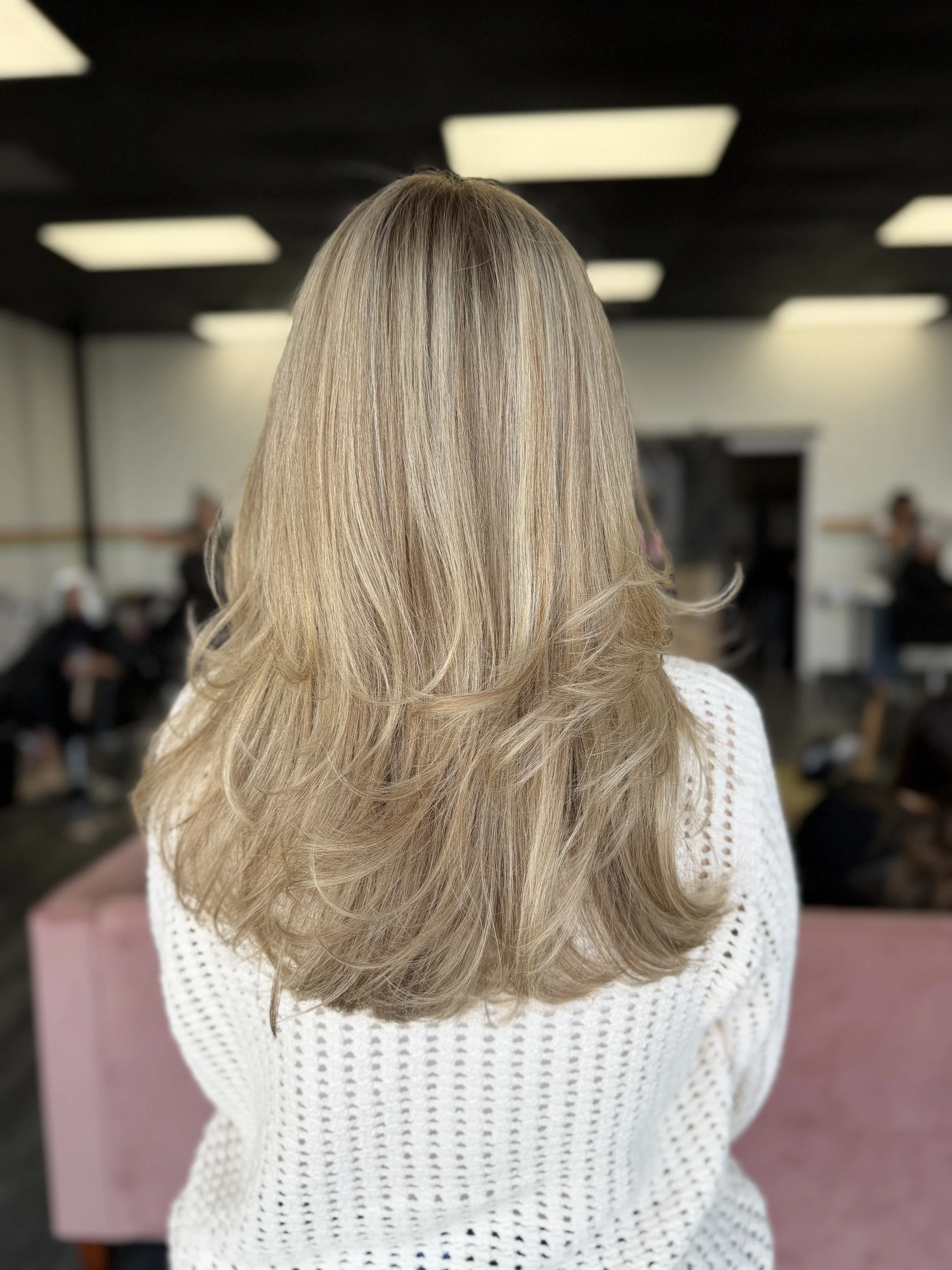 Back of a woman's head with long, layered, blonde hair, wearing a white knitted sweater, indoors with blurred background of people and ceiling lights.