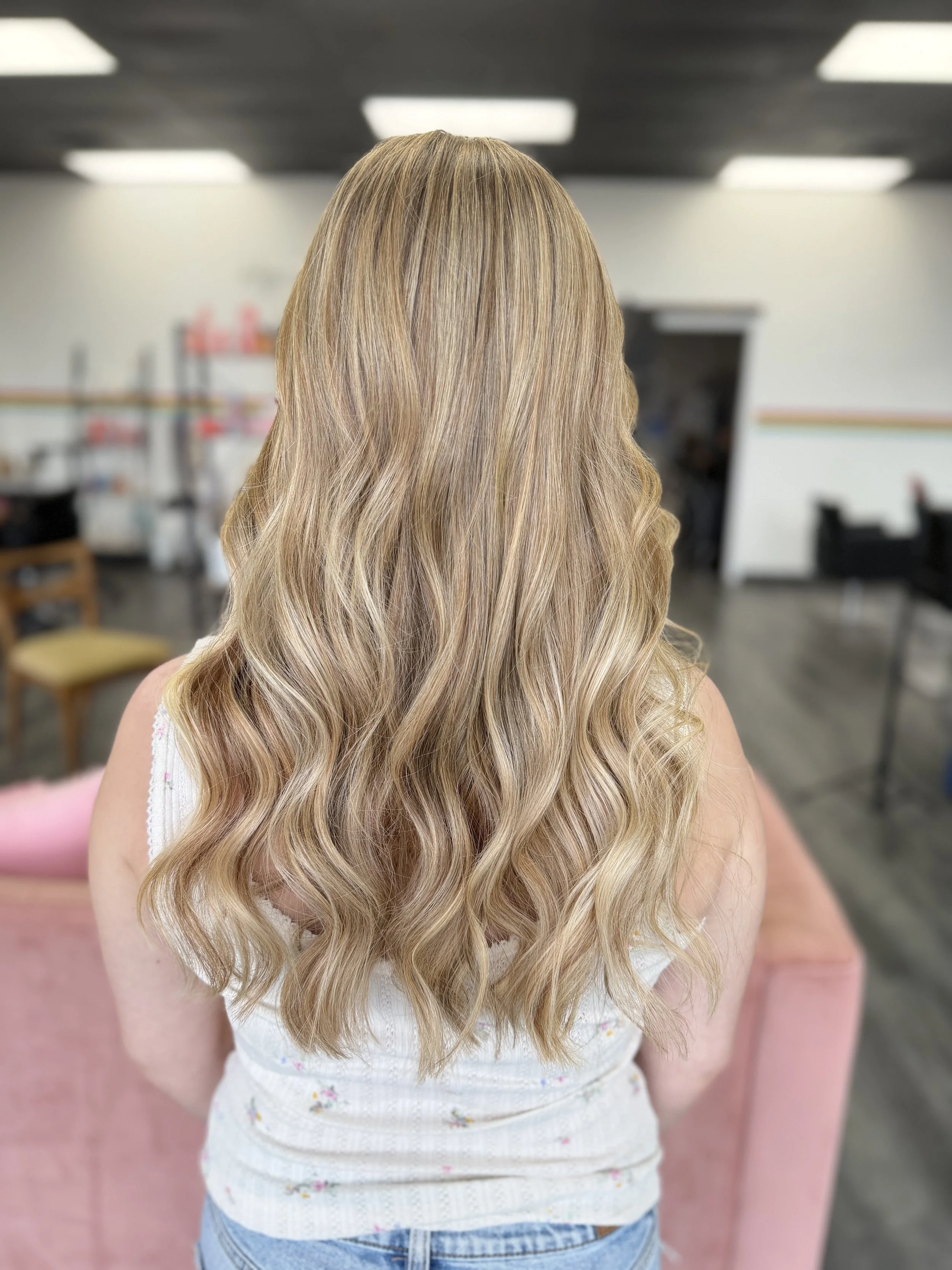 Back of a woman with long, wavy blonde hair, wearing a white top with floral patterns and blue jeans, in what appears to be a hair salon.