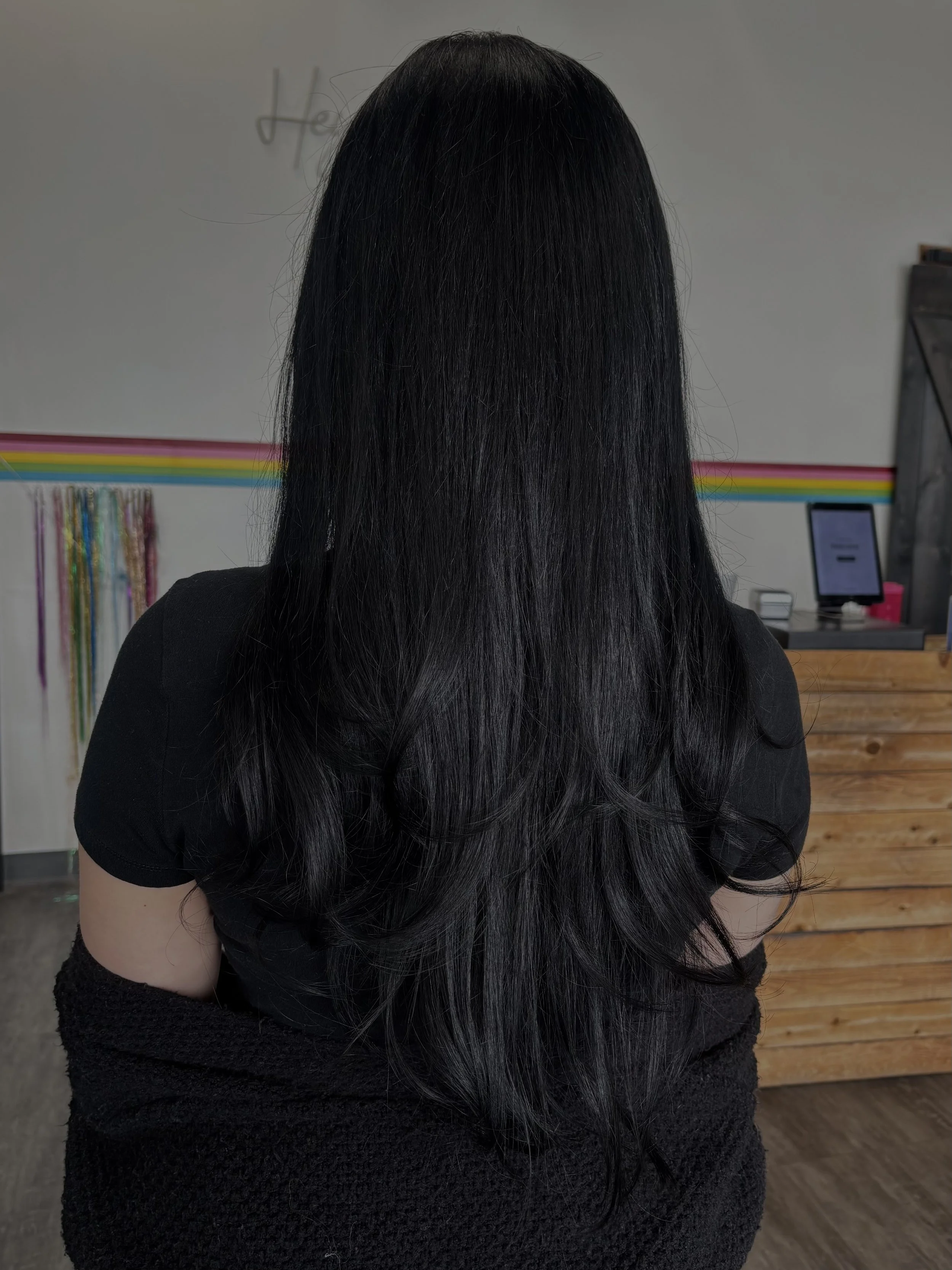 Back view of a woman with long, straight black hair, wearing a black top, standing indoors in front of a wall with colorful horizontal stripes and a countertop with a monitor and other items.