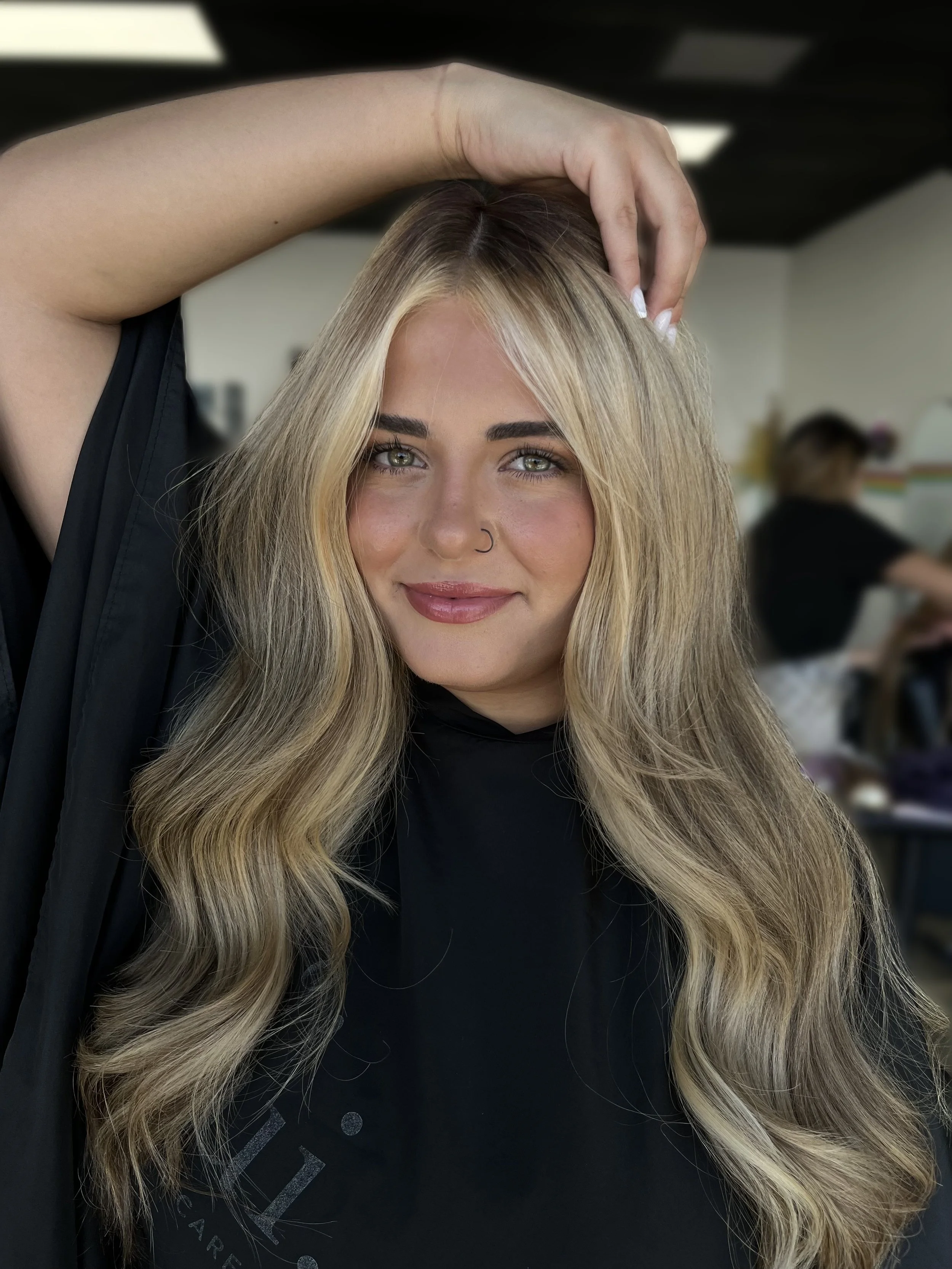 Close-up of a woman with long, wavy blonde hair, smiling, with her hand resting on her head, in a salon setting.