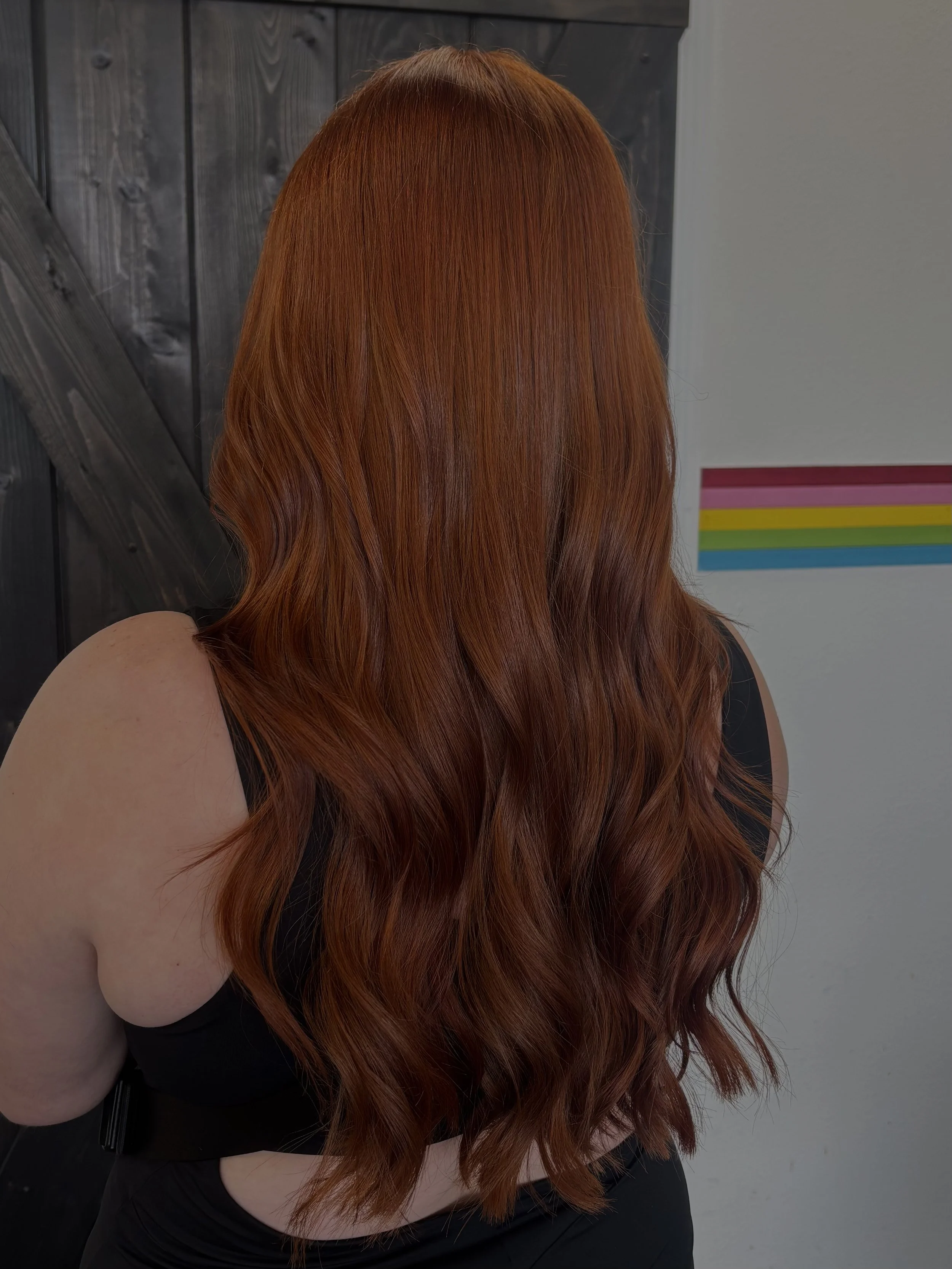 Back of a woman with long, wavy, reddish-orange hair standing against a dark wood wall on the left and a gray wall with a colorful striped poster on the right.