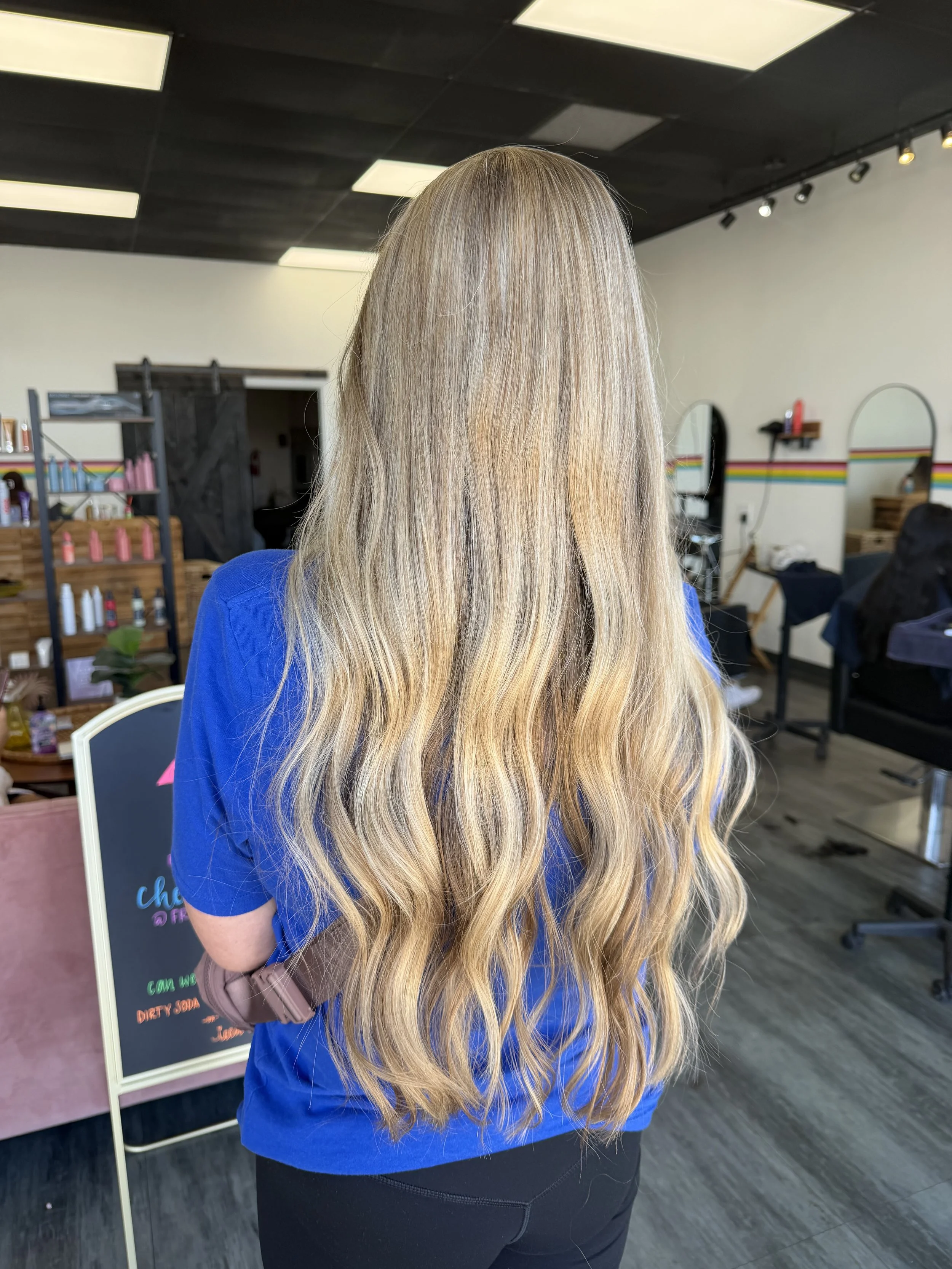 Back view of a woman with long, blonde, wavy hair, wearing a blue shirt, standing inside hair salon.