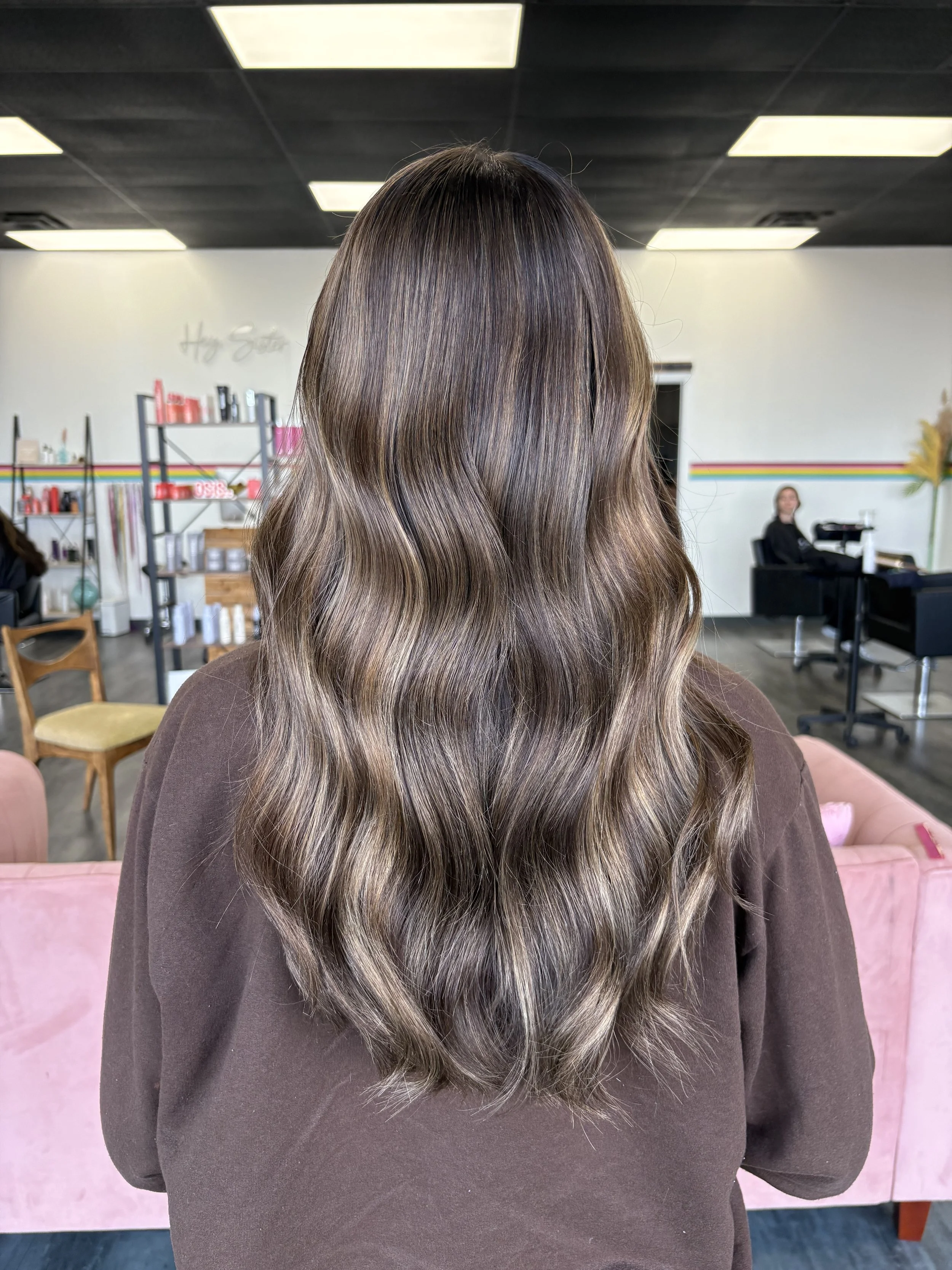 Back of a woman with long, wavy, brown hair with blonde highlights, wearing a dark brown top, sitting in a salon or studio with shelves and a person working at a desk in the background.