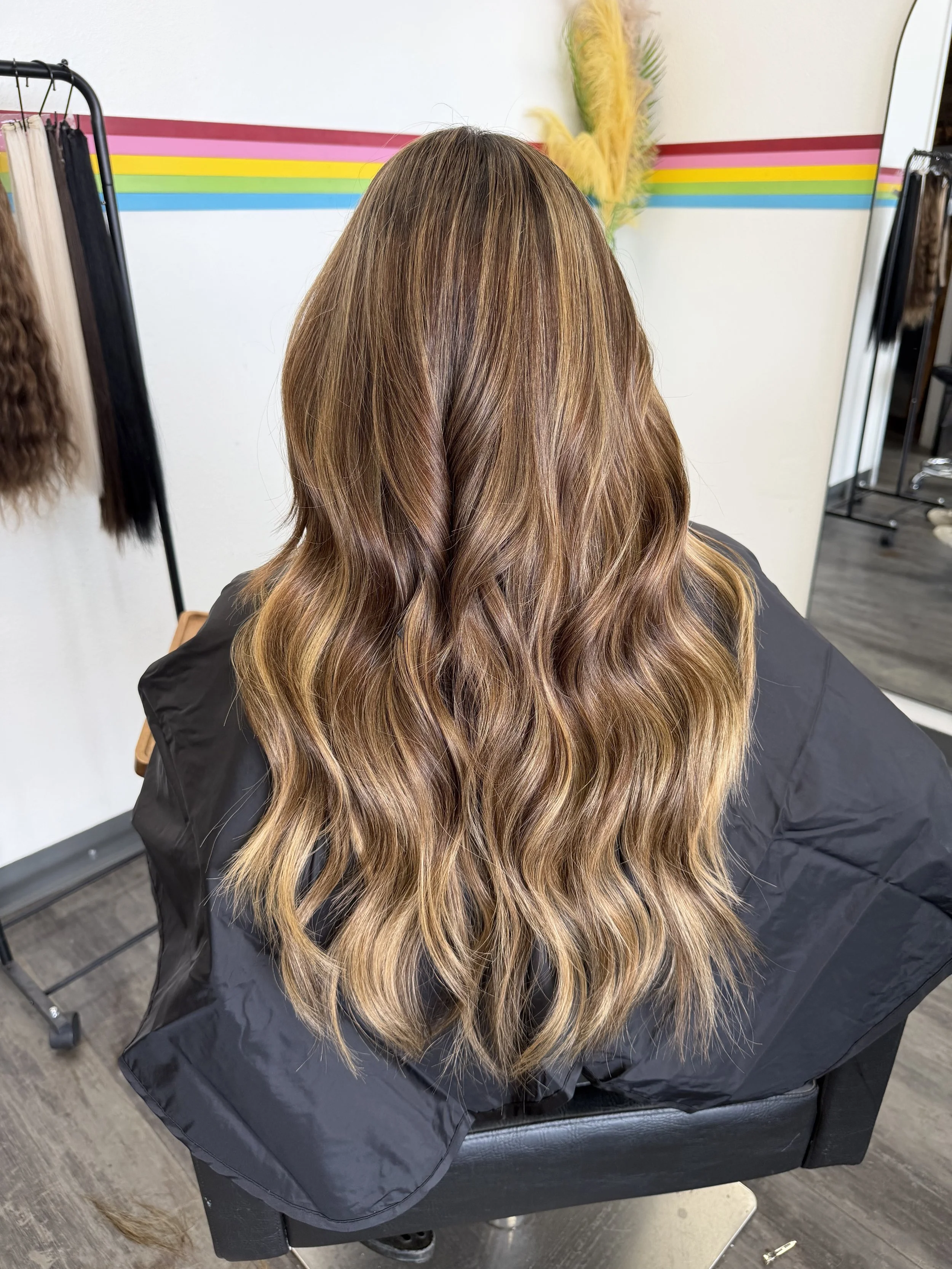 Back view of a woman with long wavy hair that has balayage highlights, seated in a salon chair with a black cape, in a hair salon with colorful striped wall and mirror.