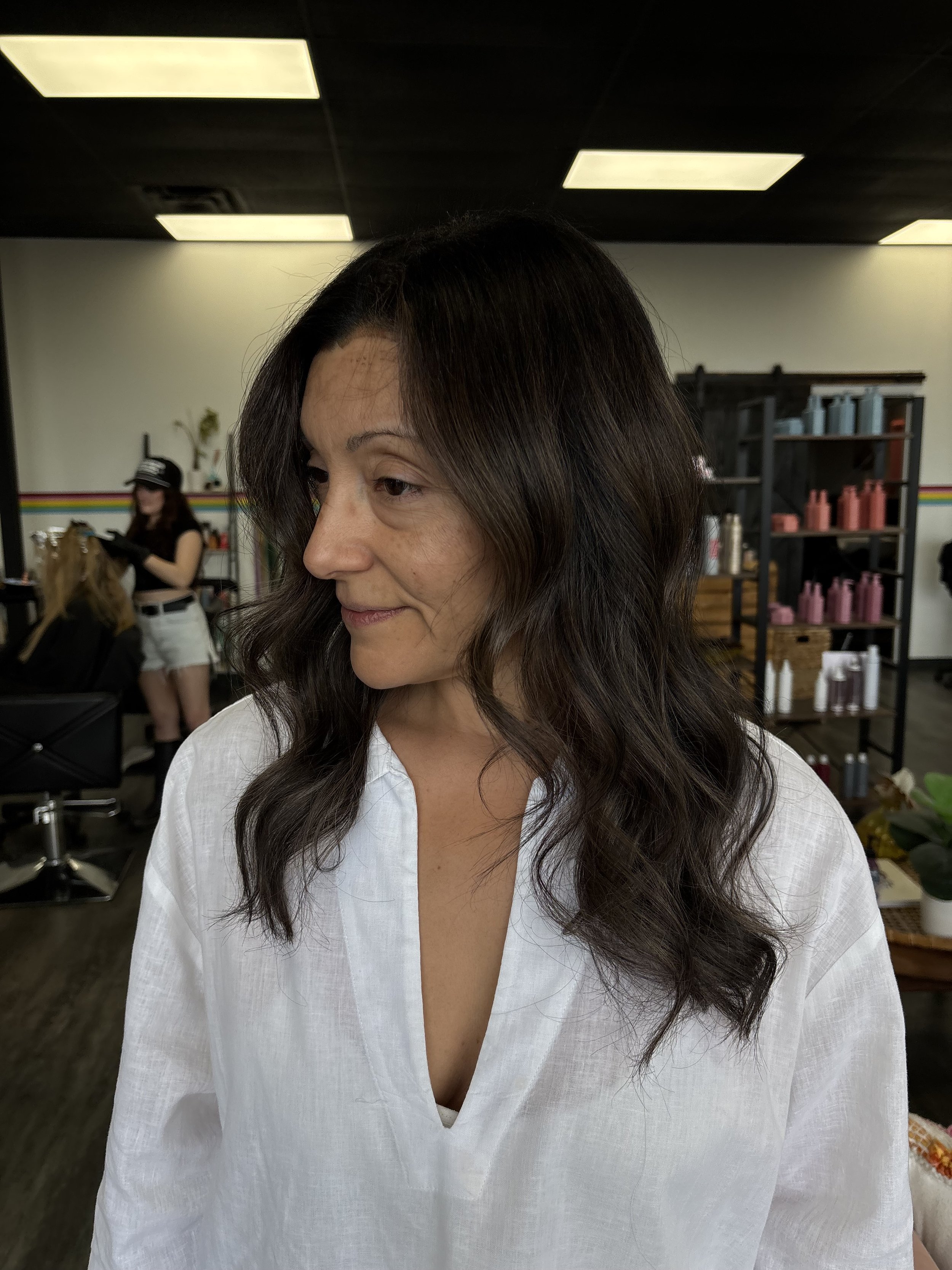 A woman with long wavy dark hair wearing a white shirt, standing in a hair salon with shelves of hair products in the background and another stylist working on a client.