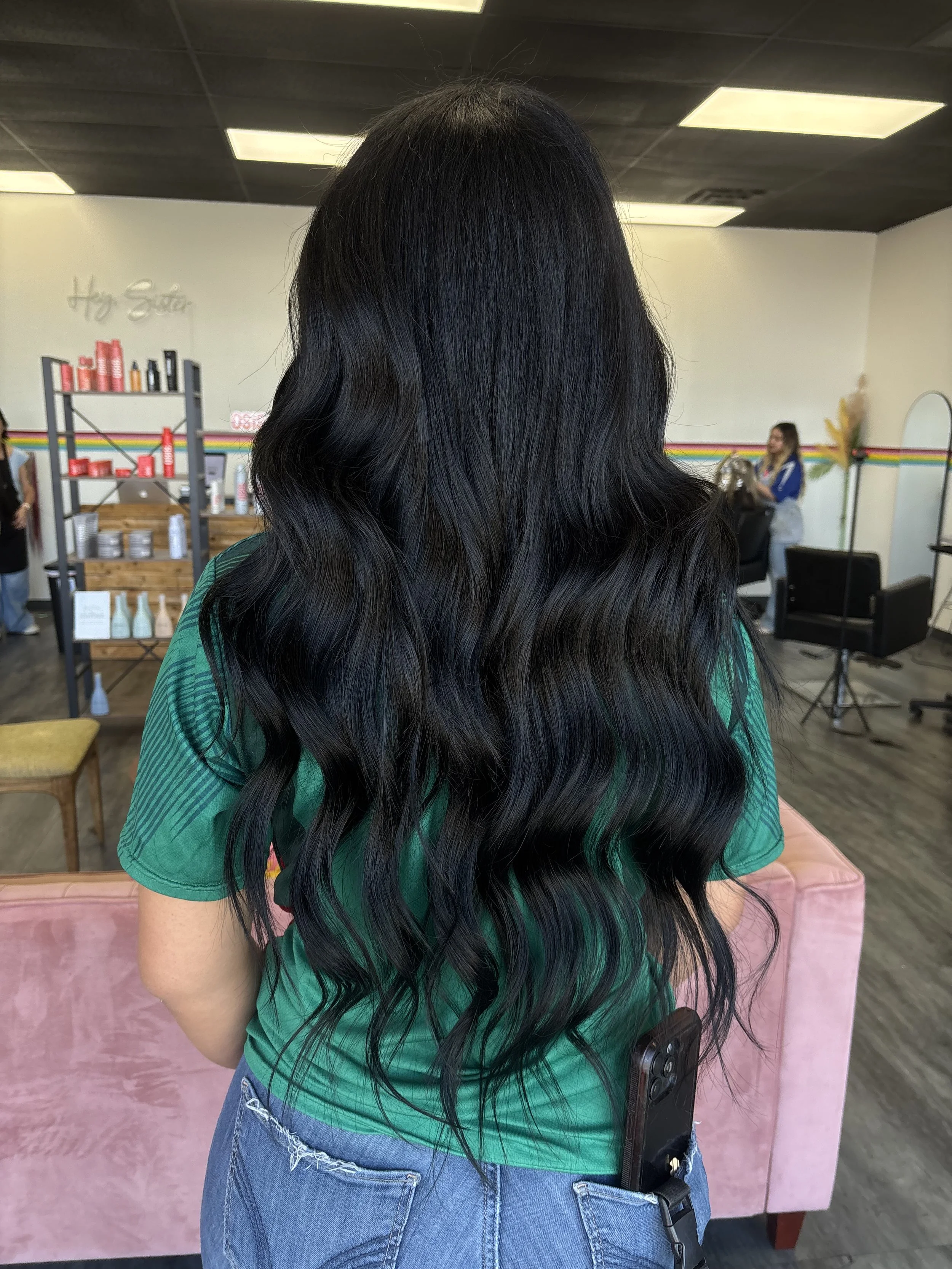 A woman with long, wavy black hair wearing a green shirt and blue jeans, sitting on a pink chair in a hair salon, with her back to the camera. The salon has shelves of hair care products and another woman working in the background.