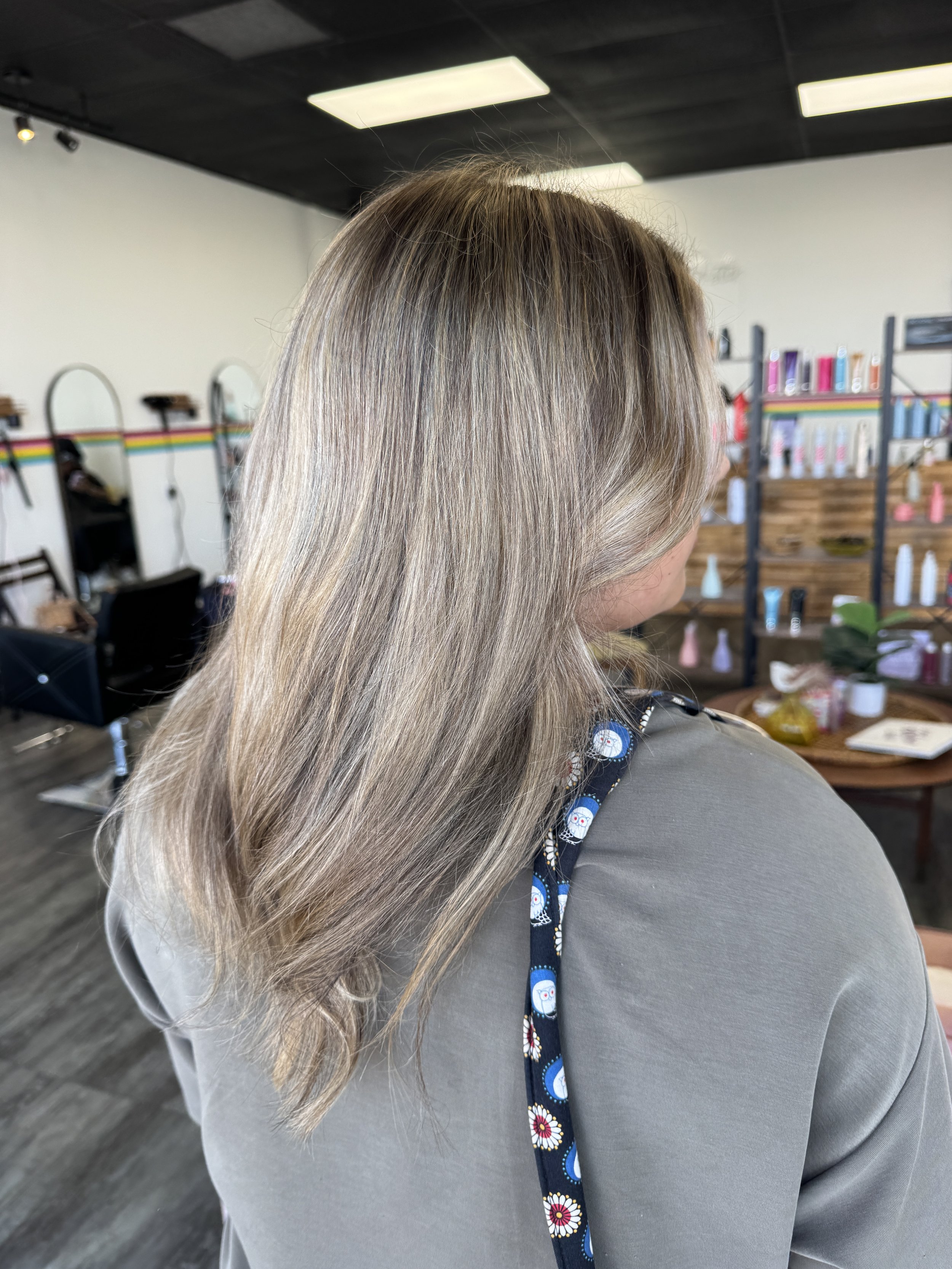 Close-up of a woman with long, straight, blonde hair wearing a gray top and a dark blue scarf with colorful flowers, inside a hair salon with shelves of hair products in the background.