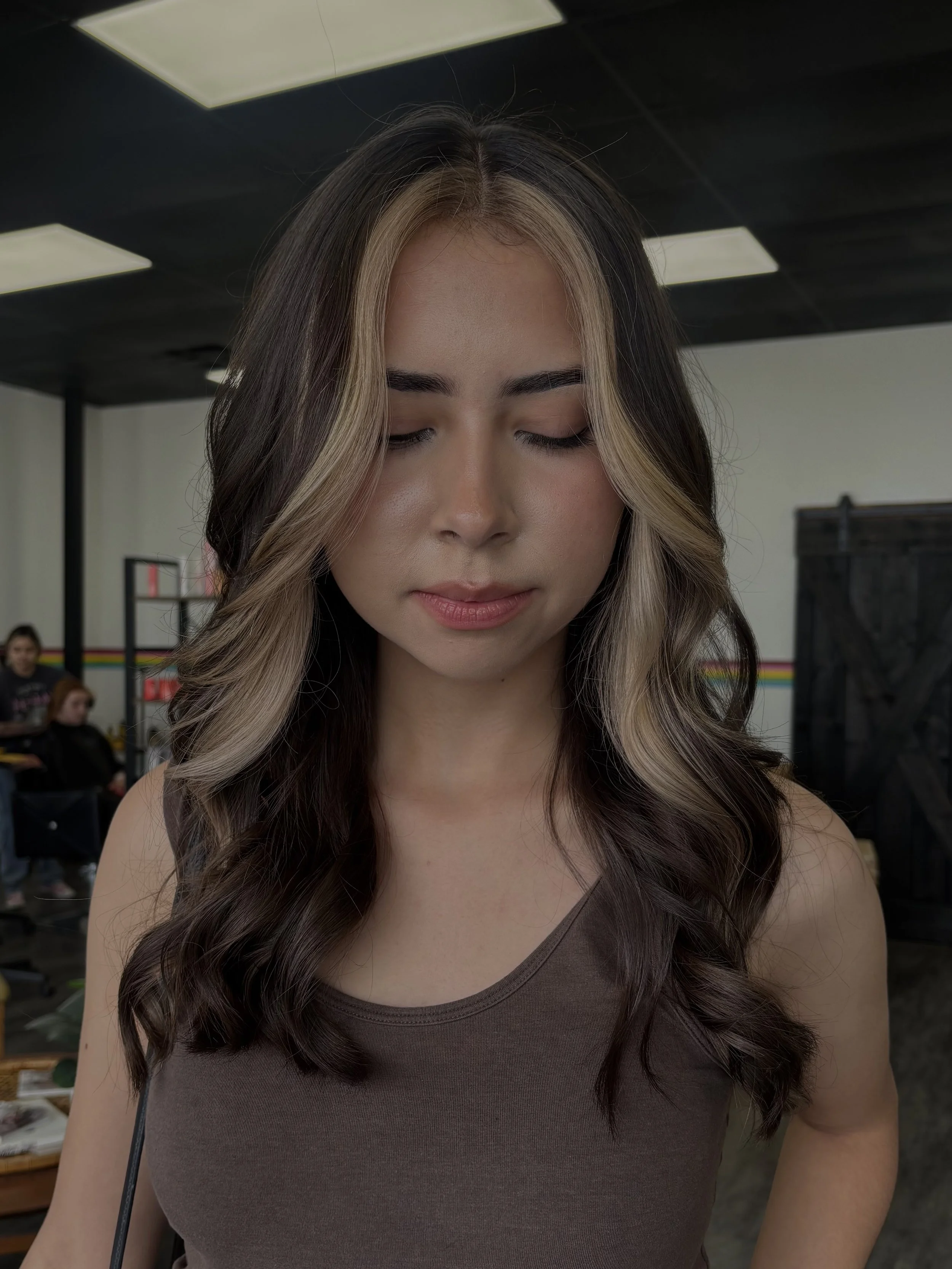 A woman with long, wavy brown hair with blonde highlights, wearing a brown sleeveless top, with a neutral expression, standing indoors.