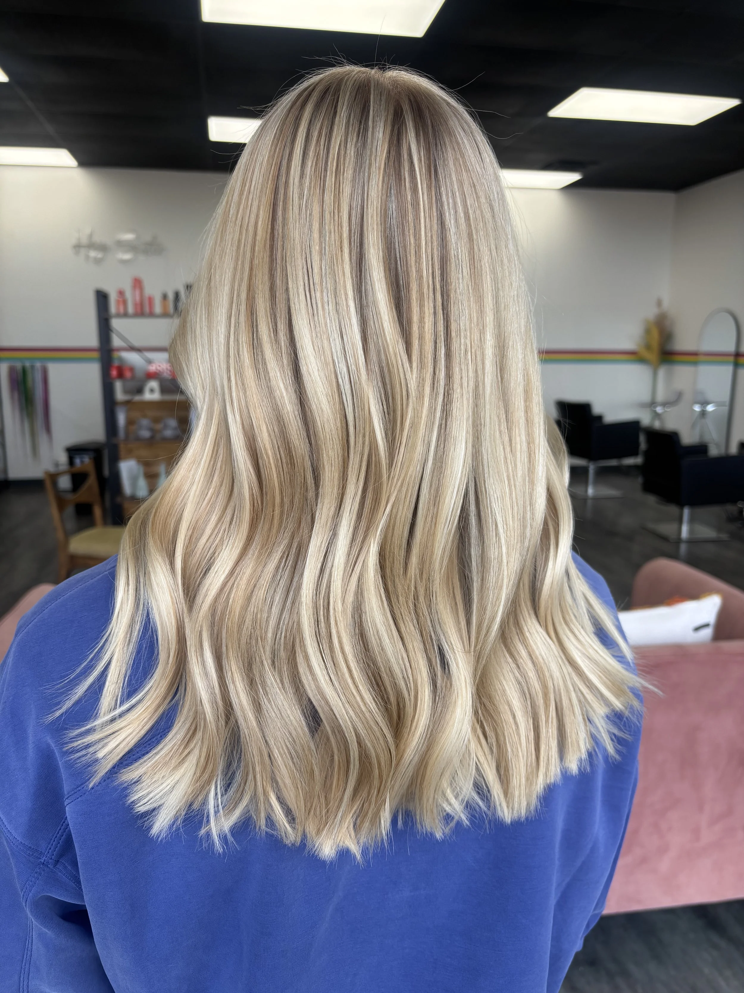 Back view of a woman with long, wavy, blonde hair wearing a blue top in a hair salon.