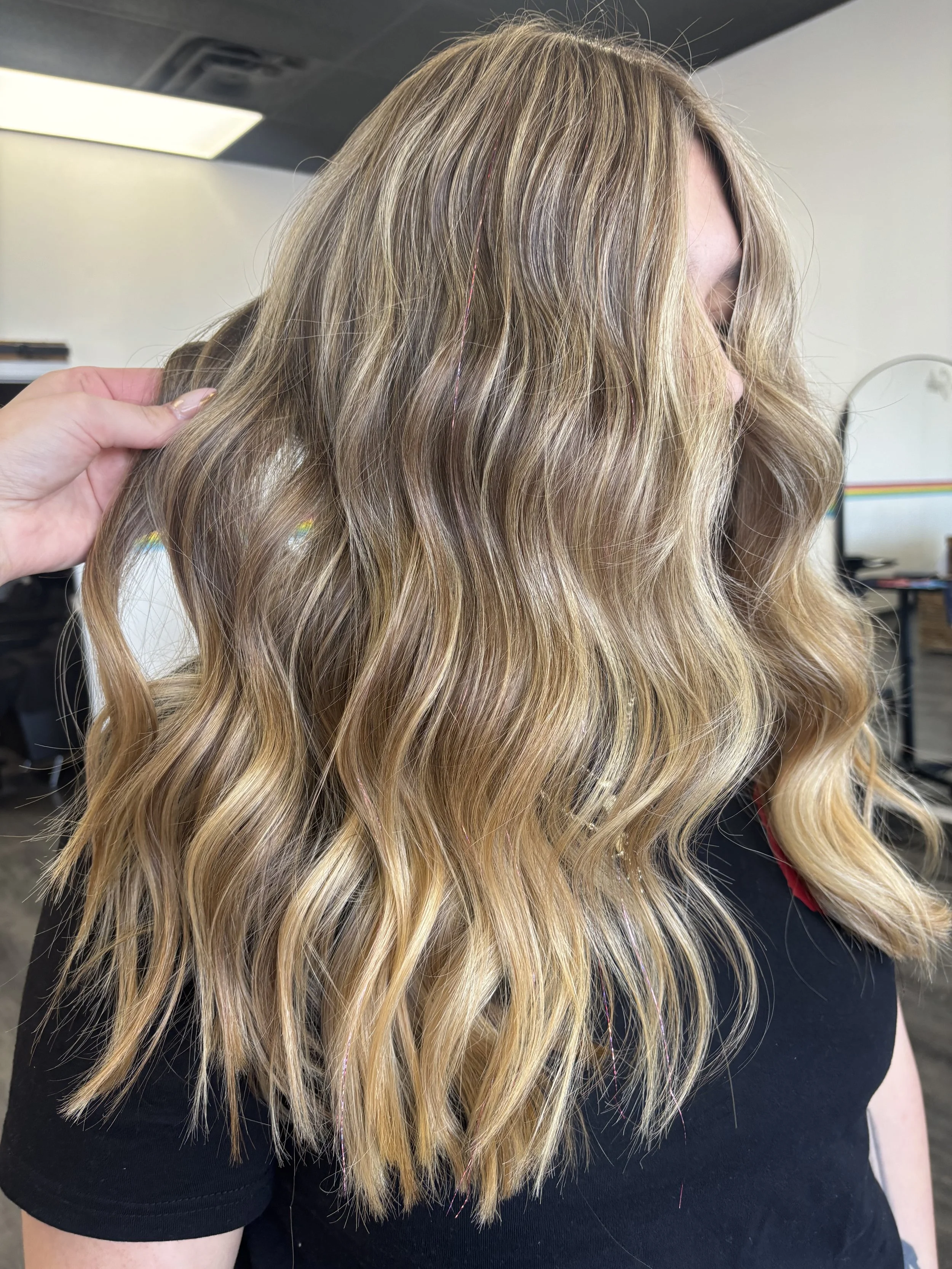 Close-up of a person's hair with blonde highlights styled in loose waves, wearing a black shirt in an indoor setting.