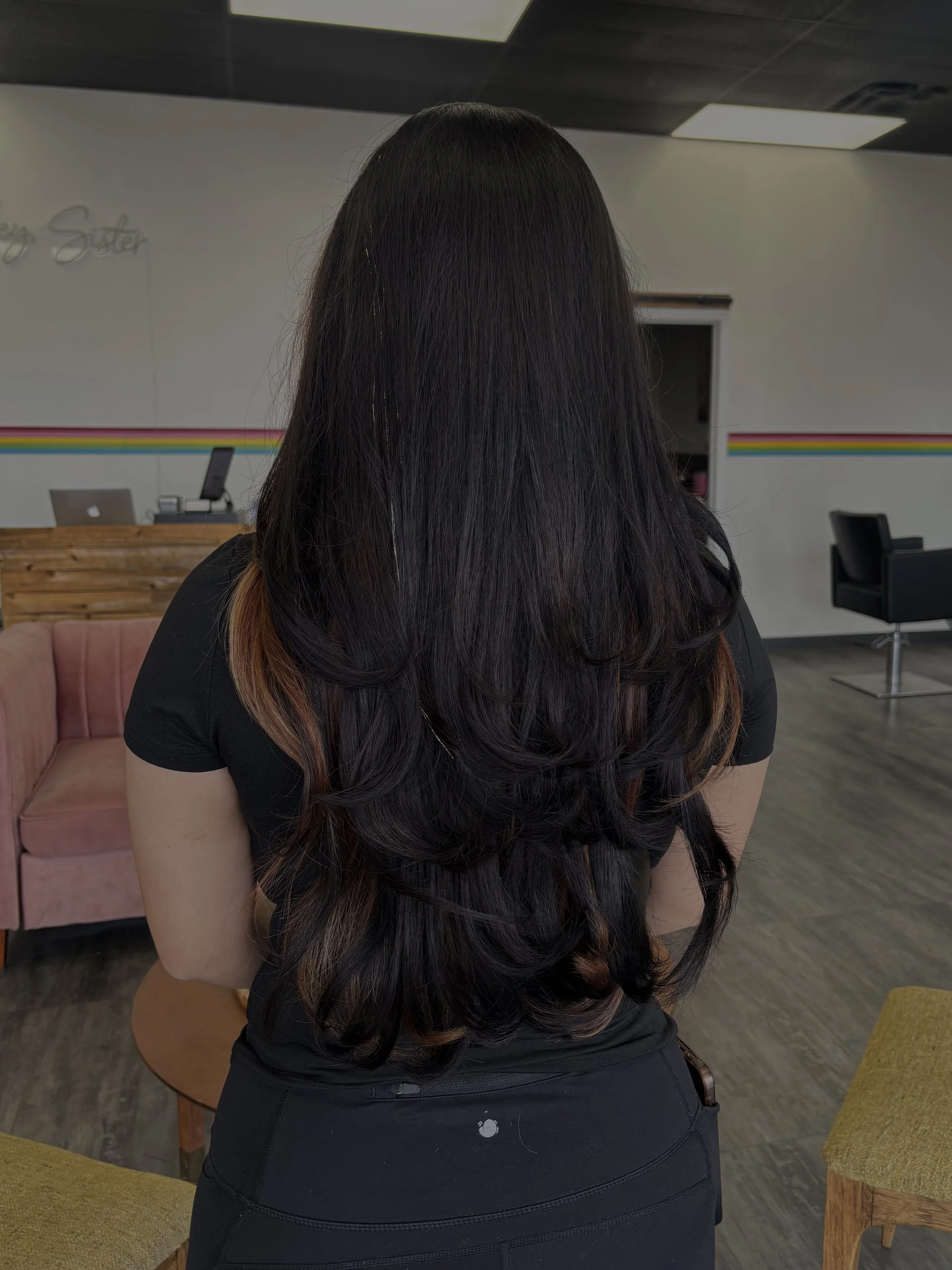 A woman with long, dark hair with brown highlights, wearing a black top and black pants, is standing indoors with her back to the camera. The room has a modern, minimalist style with a pink sofa, wooden tables, and chairs. A rainbow stripe decorates 