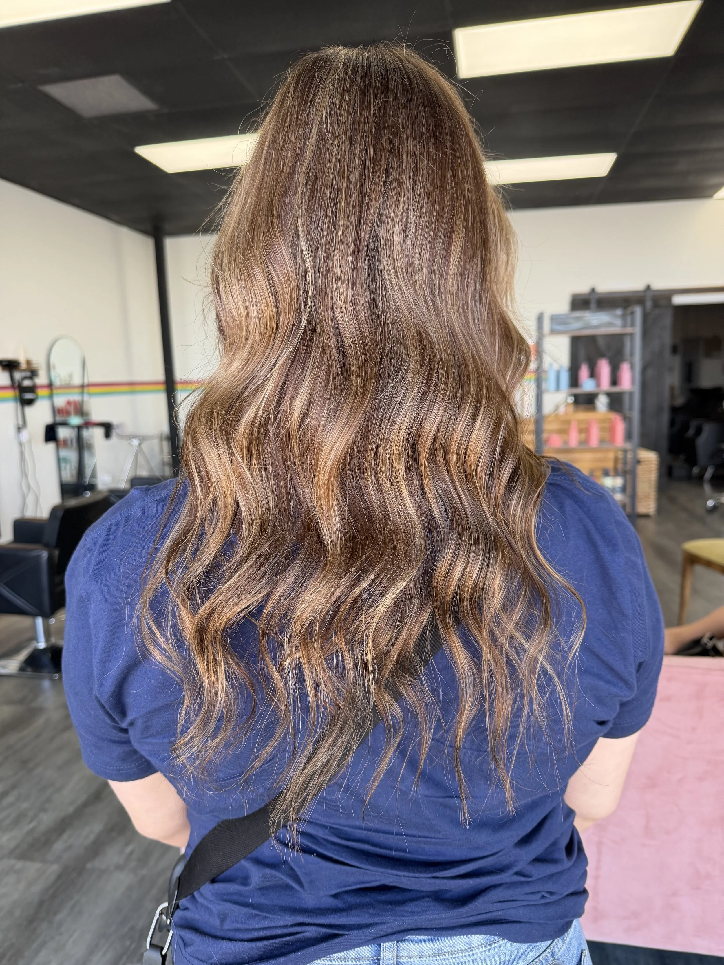 Back view of a woman with long, wavy, reddish-brown hair wearing a blue top in a hair salon.