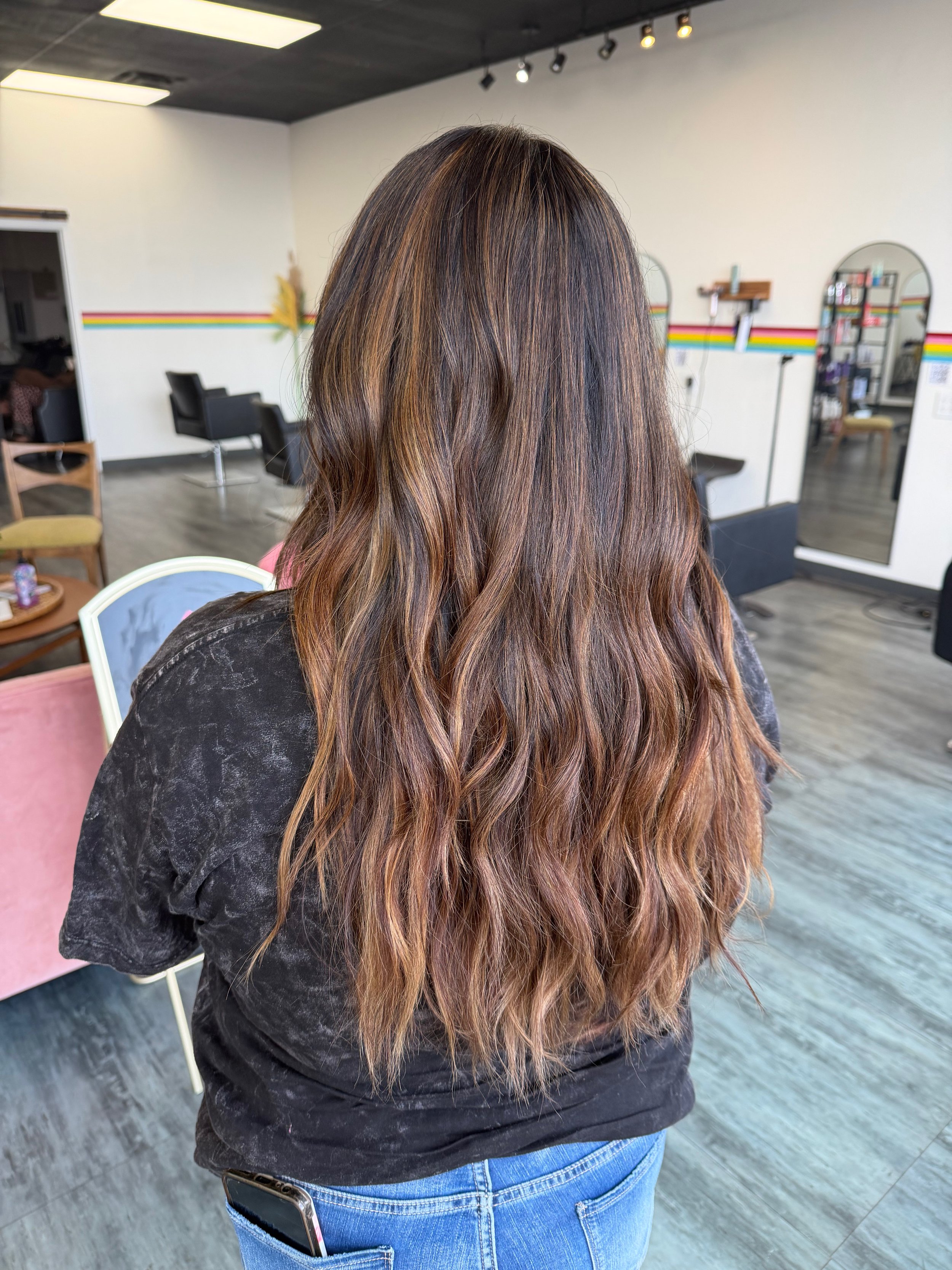 Back view of a woman with long, wavy hair that is highlighted with shades of brown and blonde, standing inside a hair salon.
