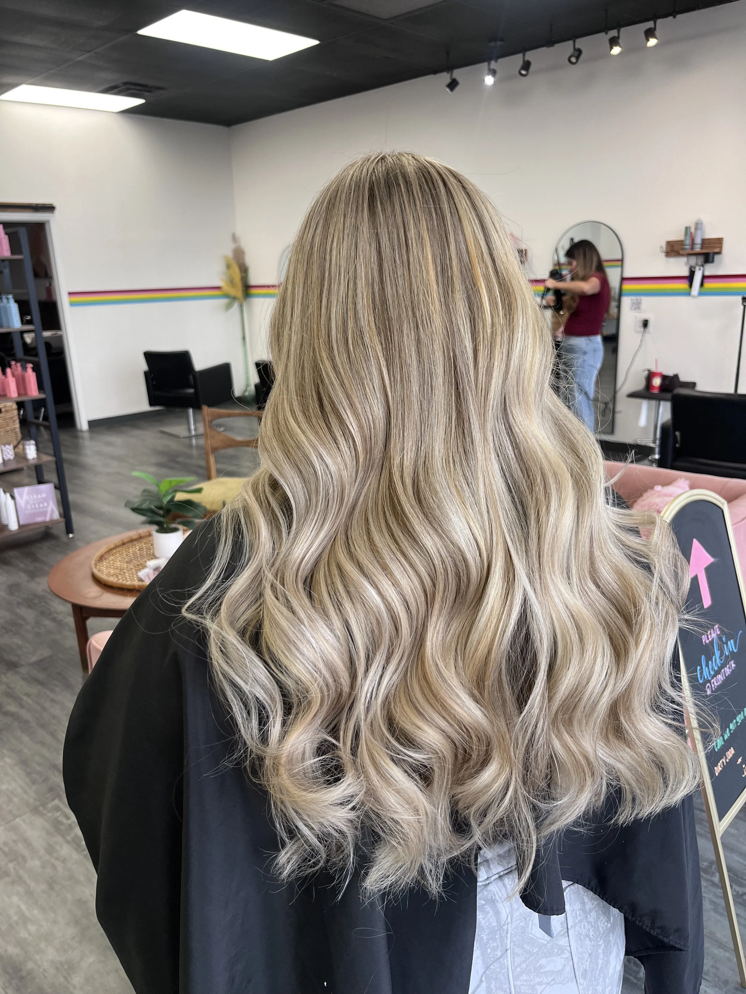 Back view of a woman with long, wavy blonde hair sitting in a salon. She is wearing a black cape, and the salon has white walls with colorful horizontal stripes, black chairs, and salon shelves.