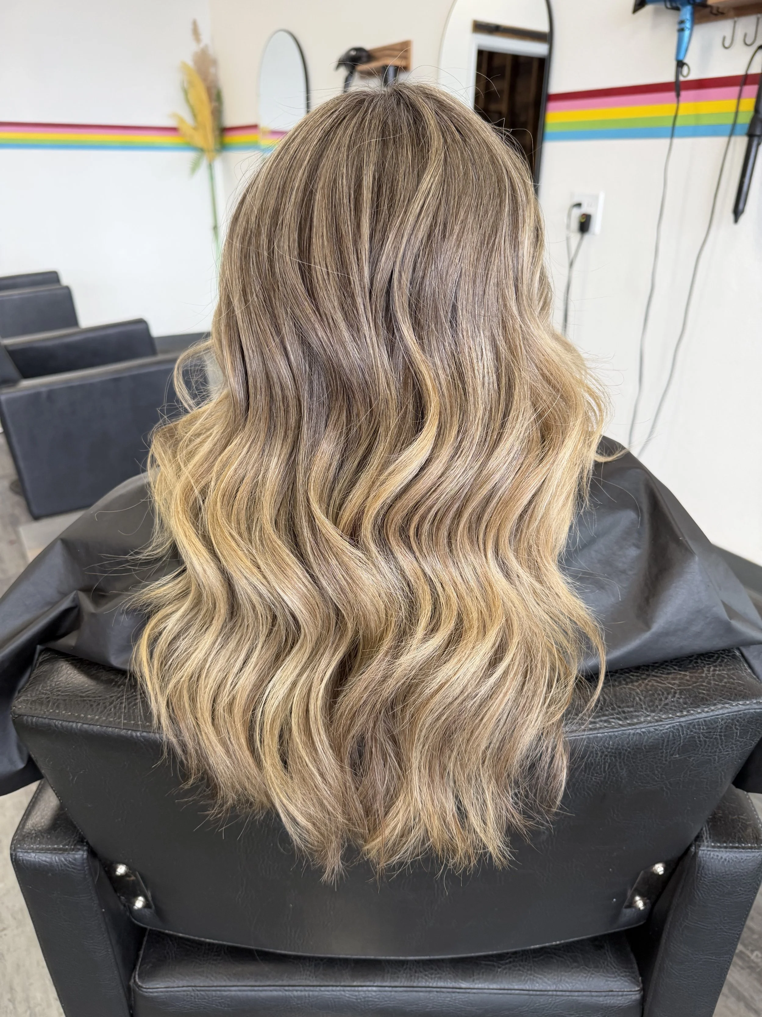 Back of a woman with long blonde wavy hair sitting in a salon chair, with mirrors and salon tools visible on the wall behind her.