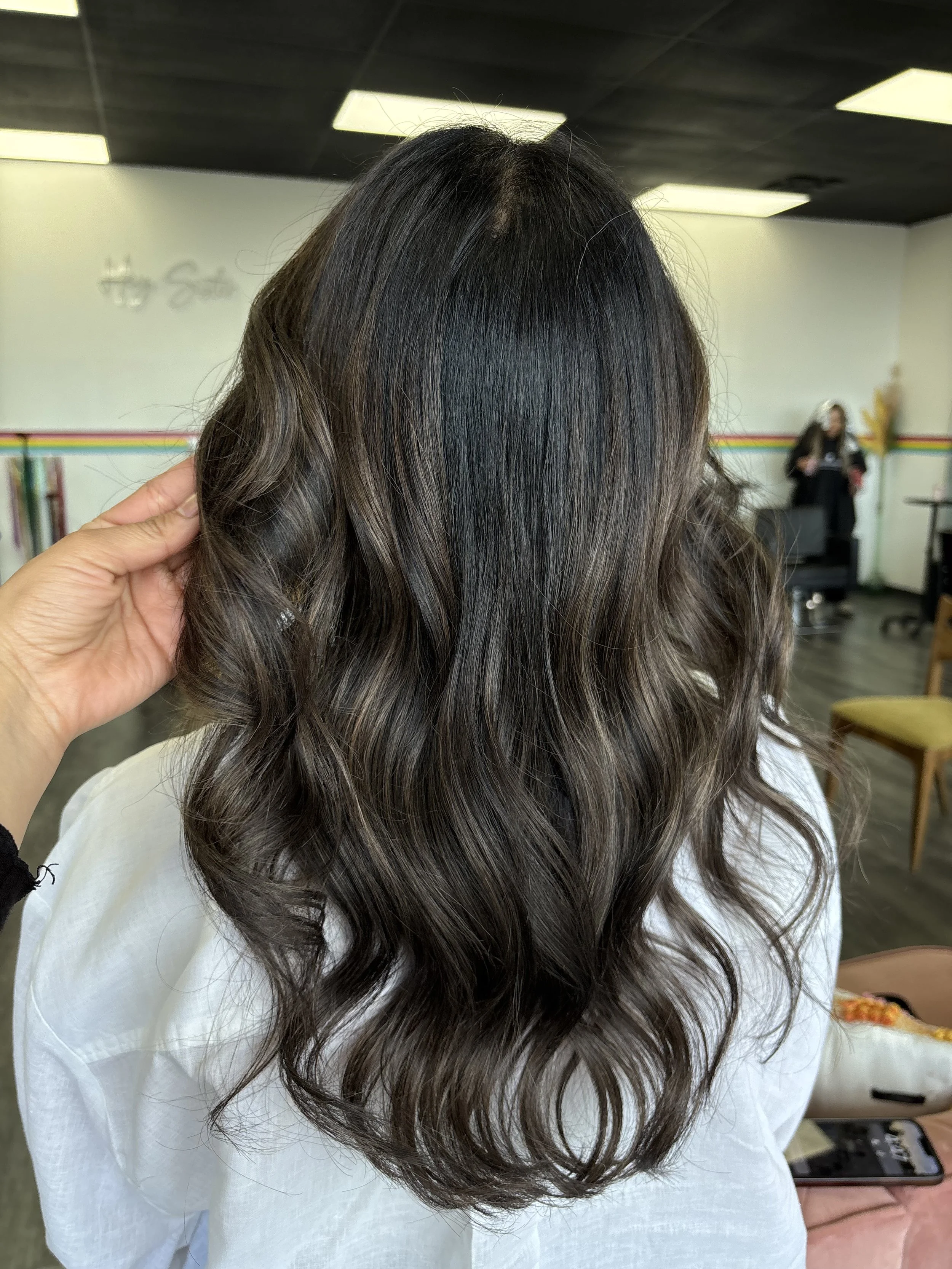 Interior of a salon with a woman sitting, hairdresser showing her long, wavy, brown hair with highlights, in front of a mirror, with another person viewing a tablet in the background.