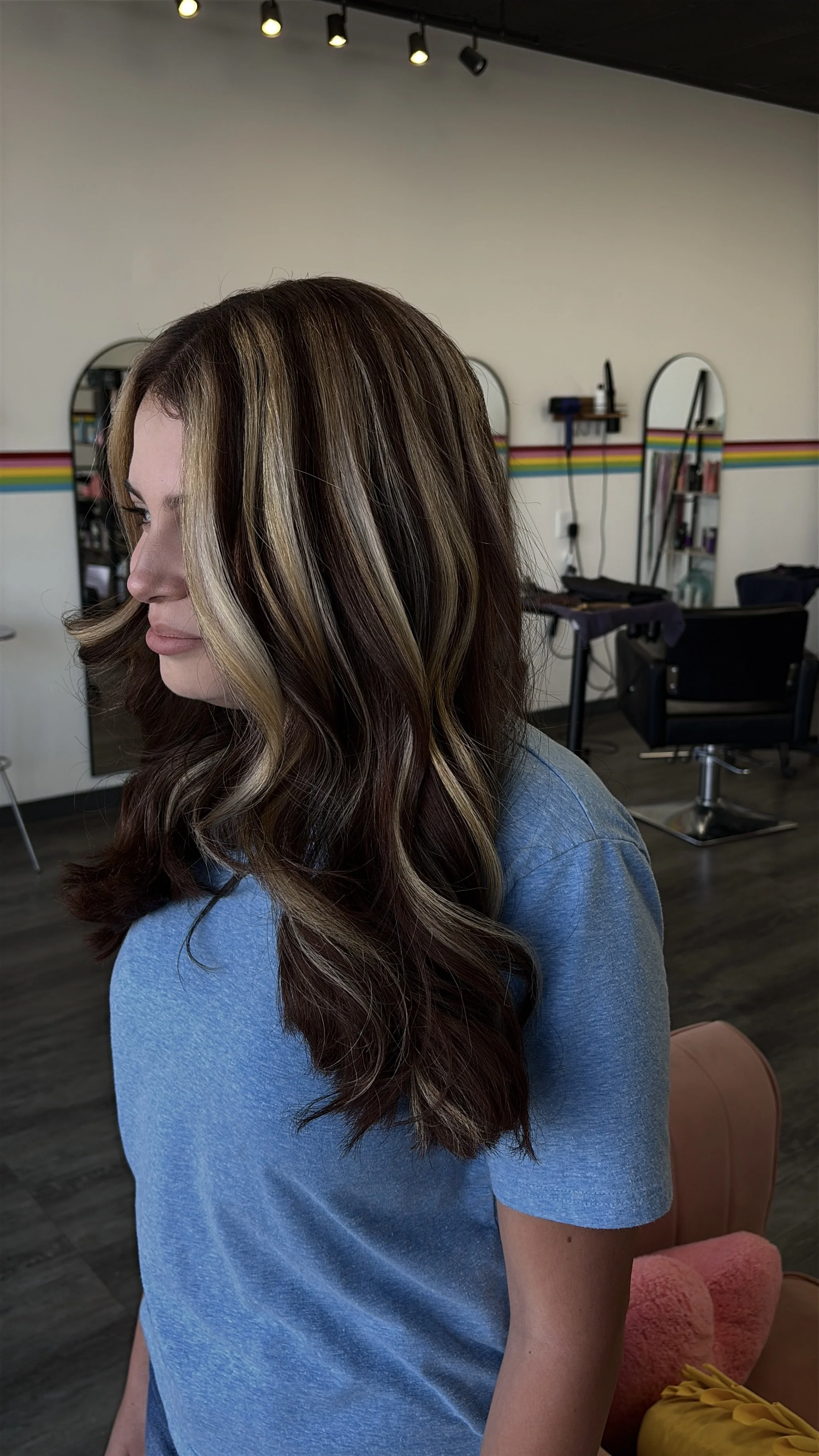 A woman with long, wavy hair featuring caramel highlights, wearing a blue t-shirt, sitting in a hair salon with mirrors and salon equipment in the background.