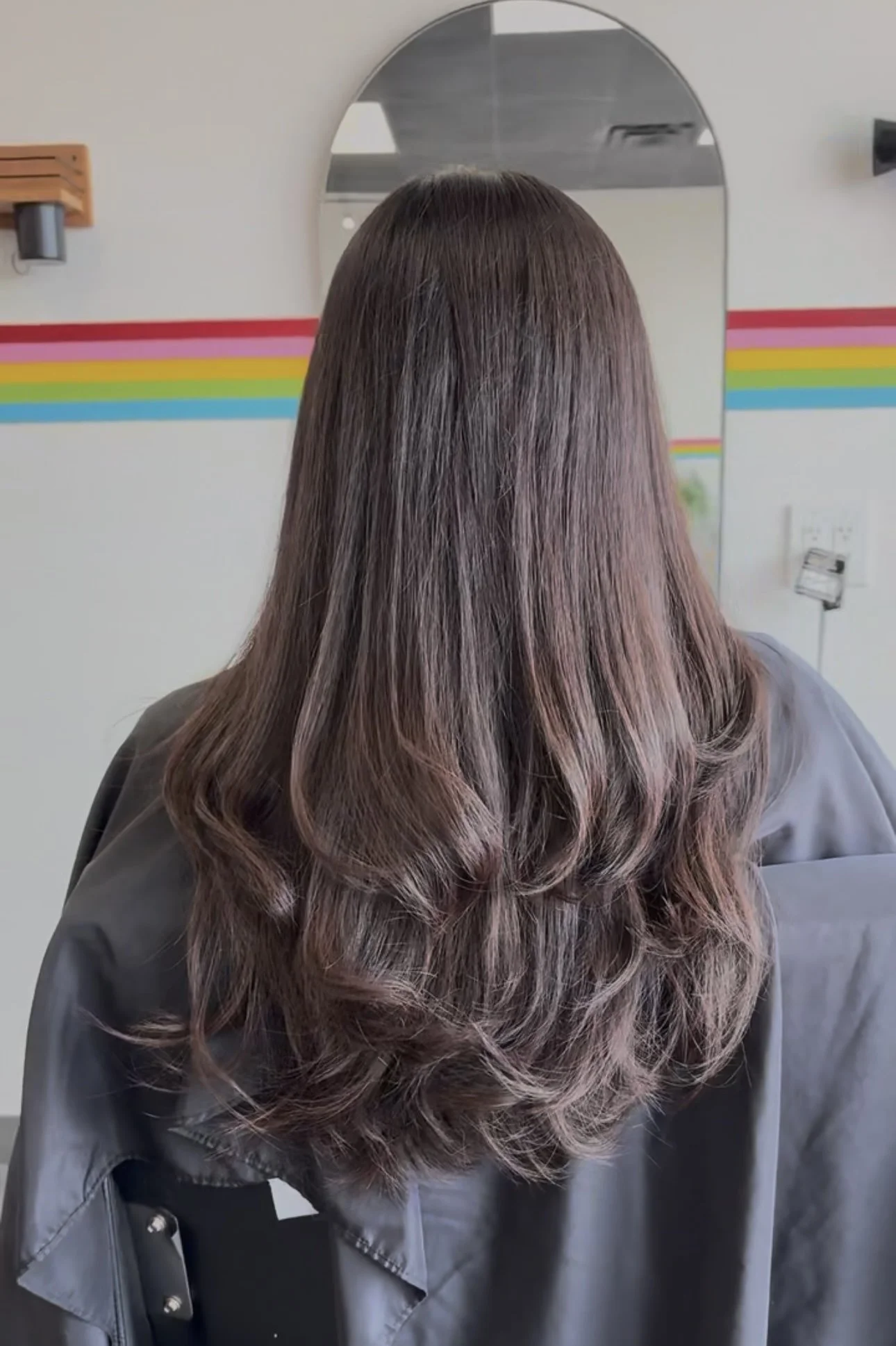 A woman with long, wavy, brown hair with highlights, wearing a gray jacket, standing in front of a mirror in a room with rainbow-colored stripes on the wall.