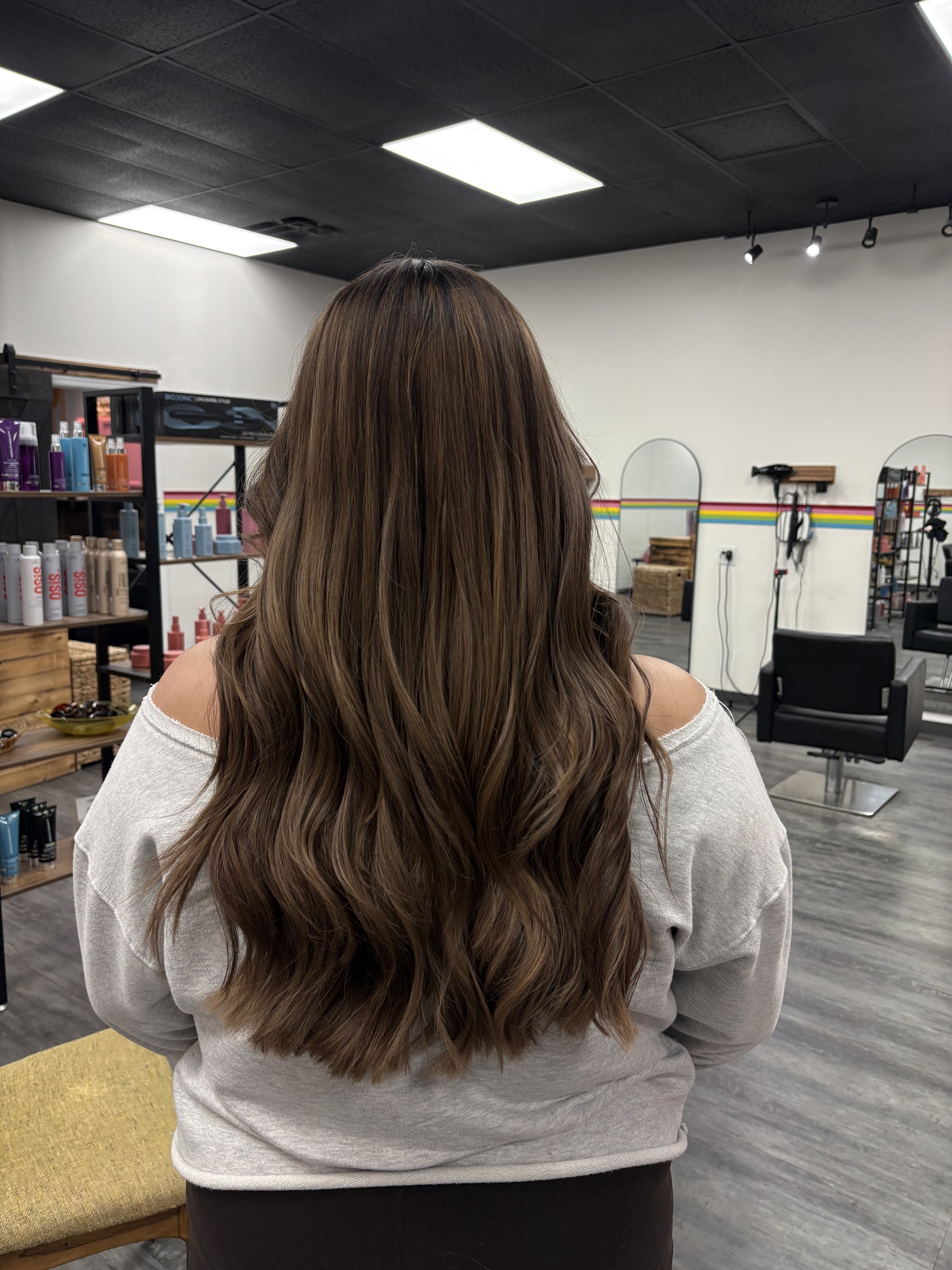 Back view of a woman with long, wavy brown hair in a hair salon, wearing a white off-shoulder top and dark pants. Salon equipment and products visible in the background.
