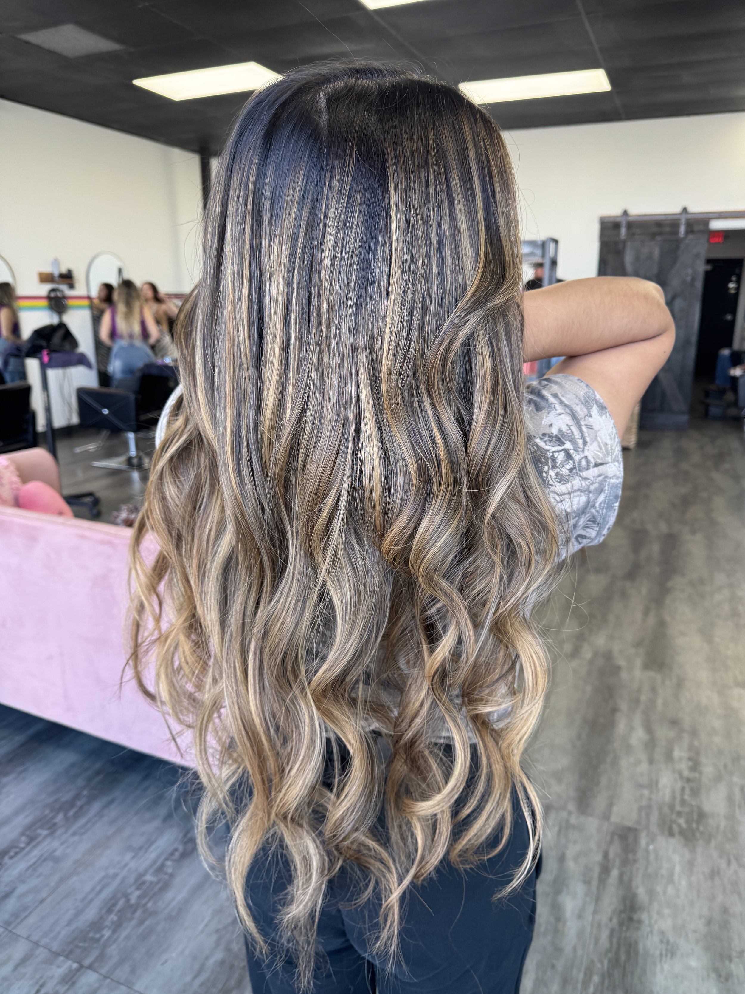 A woman with long, wavy hair showcasing caramel blonde highlights in a salon setting.