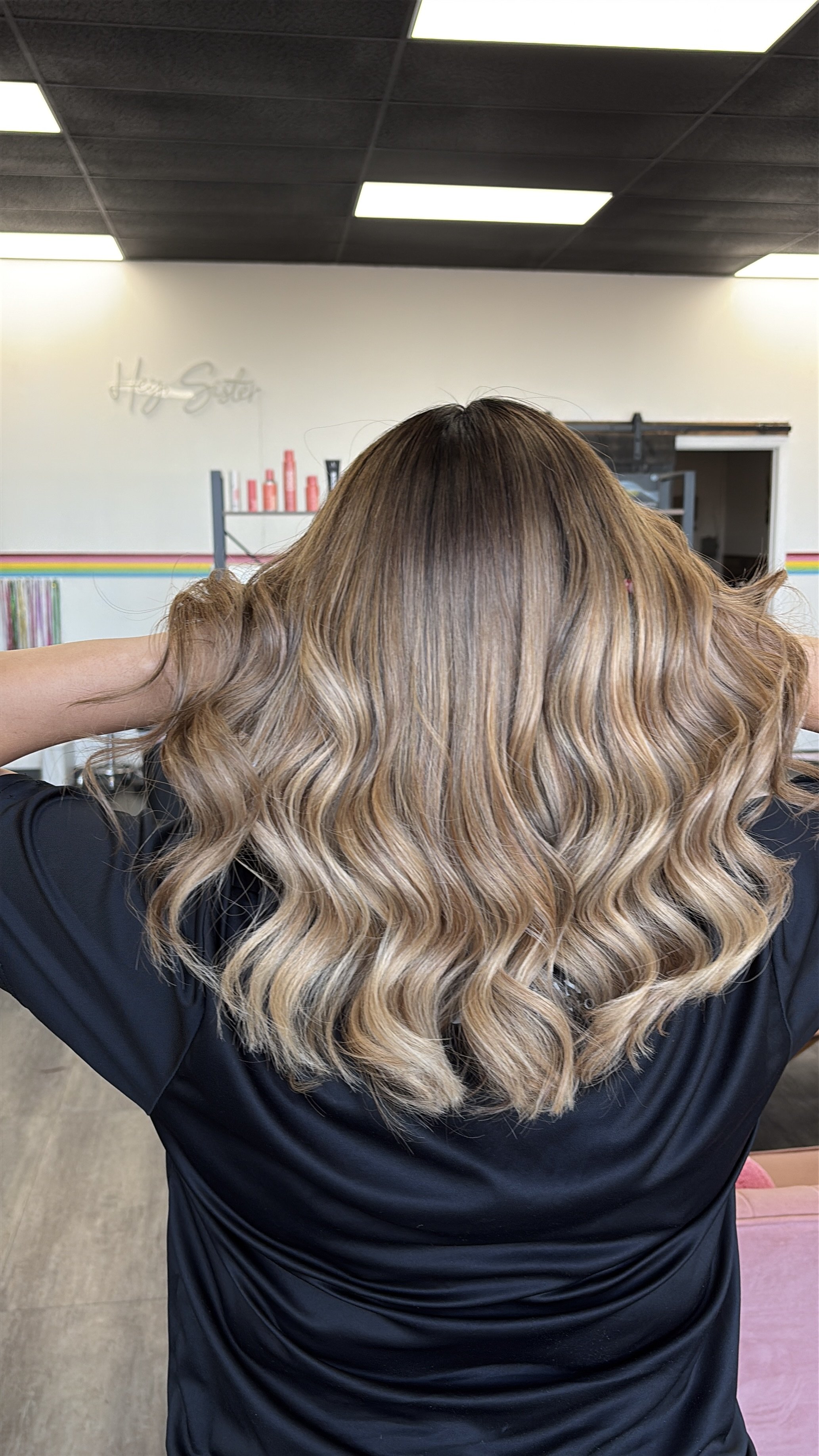 A woman with long, wavy, blonde hair with darker roots in a salon, facing away, with her hands on her head.