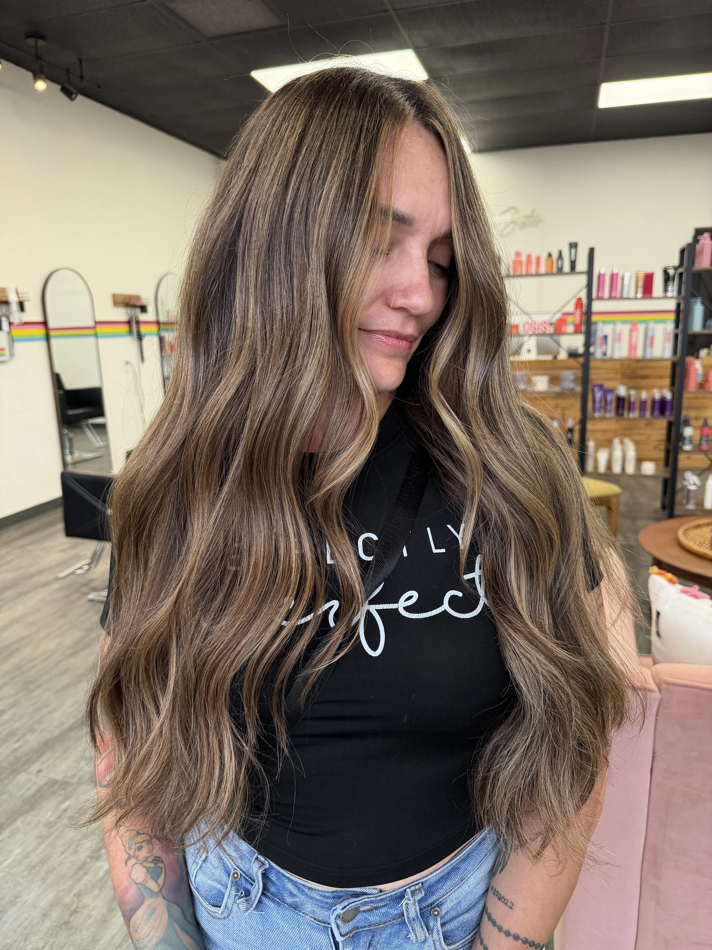 Close-up of a woman with long, wavy hair with blonde highlights, wearing a black t-shirt and light blue jeans, standing in a salon with shelves of hair products in the background.