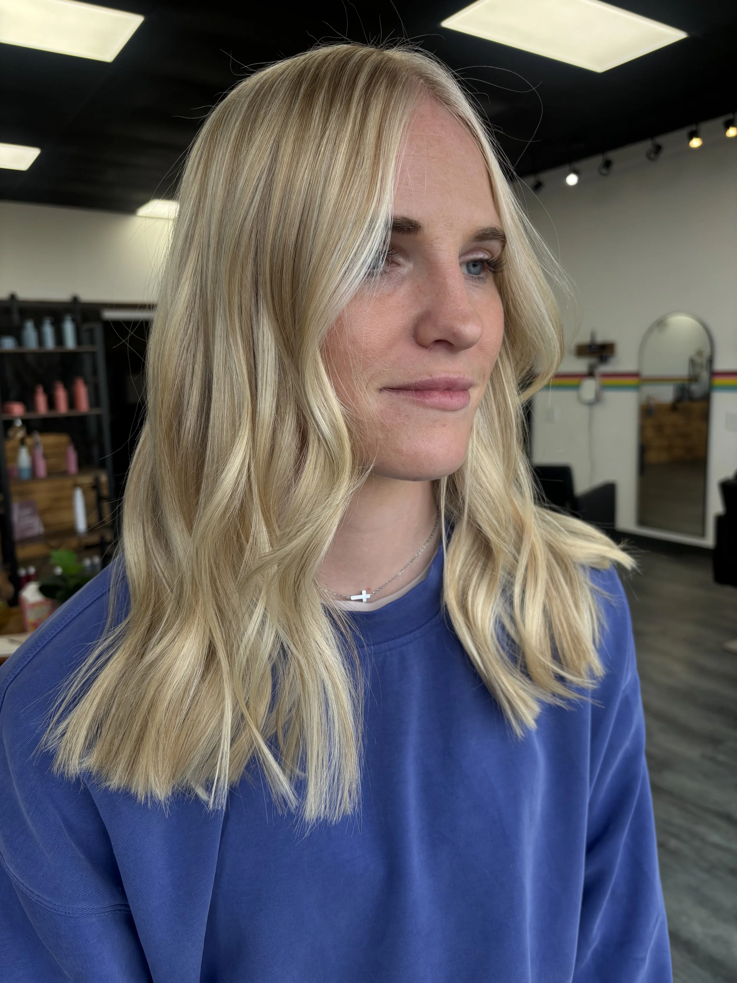 Close-up of a young woman with long blonde hair and blue eyes, wearing a blue shirt and a silver cross necklace, in a hair salon.